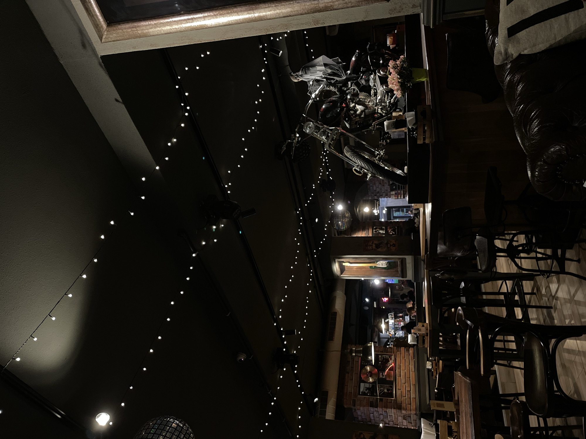 The interior of a dimly lit bar or restaurant in Veliko Tarnovo, Bulgaria is visible. The scene features hanging string lights, a motorcycle suspended from the ceiling, and a bar area with seating. Seating consists of black chairs at the tables. Walls are covered with decorative items, including music memorabilia and brick elements. A person is visible in the background at the bar.