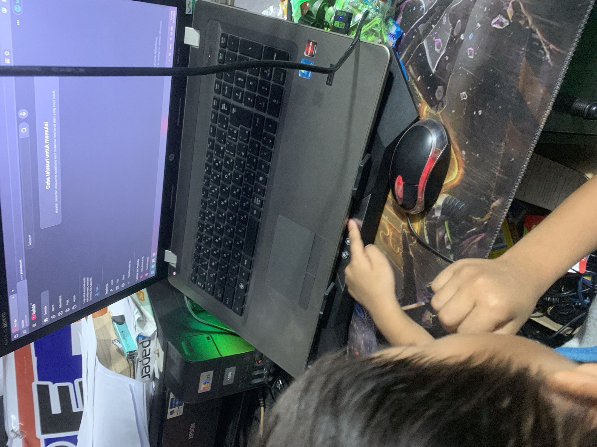 The image depicts an indoor scene, likely a home or office, with a young person interacting with a laptop. The laptop screen displays a YouTube interface, suggesting the person is browsing or watching videos. A black mouse with red accents is positioned on a patterned mousepad. The background includes a printer and stacks of paper, hinting at a cluttered workspace. The context of Sungai Meriam, Indonesia, is provided as the location. The overall impression is of a casual, everyday activity in a personal setting.