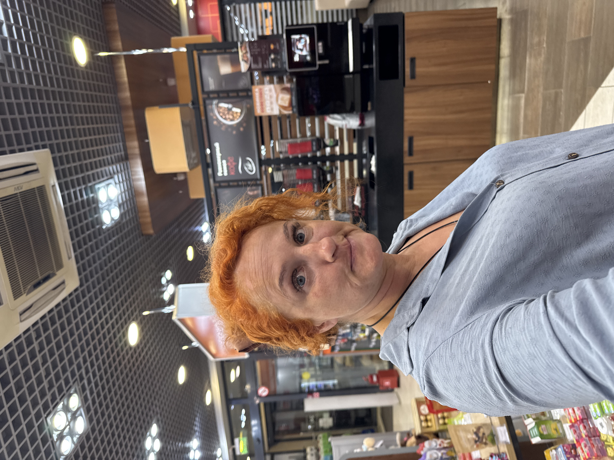 A selfie-style medium shot captures a woman with bright orange, curly hair and fair skin looking slightly upwards and to her right with a quizzical expression. She is wearing a light blue, collared, button-up shirt with a textured pattern and a black cord necklace. The background appears to be an interior retail space, possibly a convenience store or cafe, with a dark tiled ceiling with recessed lighting and air conditioning units. To her right, there is a coffee station with various dispensers and a black machine with a screen displaying an image. Shelving units stocked with products are visible behind the coffee station and to the left of the frame. The overall lighting is bright, indicative of an indoor commercial setting. There is no specific indication of the location being in the city of Можга, Russia, beyond the prompt.