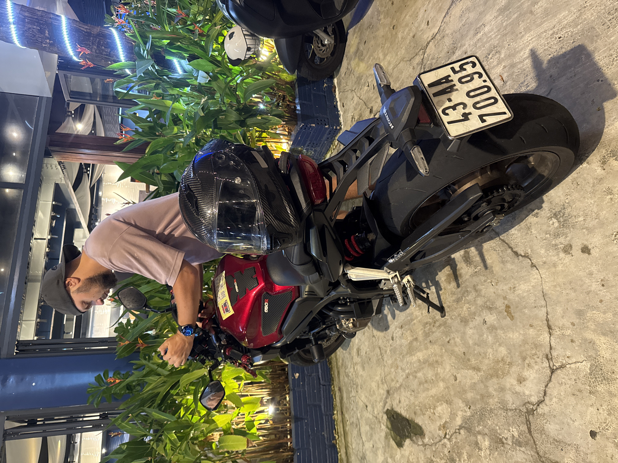 The image presents a man interacting with a red Honda motorcycle in what appears to be a public space in Đà Nẵng, Vietnam. The man, wearing a gray cap and a light pink t-shirt, is positioned near the handlebars of the motorcycle. The motorcycle has a black helmet placed on the seat. A Vietnamese license plate reading "43-A4 700.95" is affixed to the rear of the motorcycle. Nearby, there are various plants and some artificial lighting. Another motorcycle is partially visible in the background. The ground is paved with a concrete-like material.