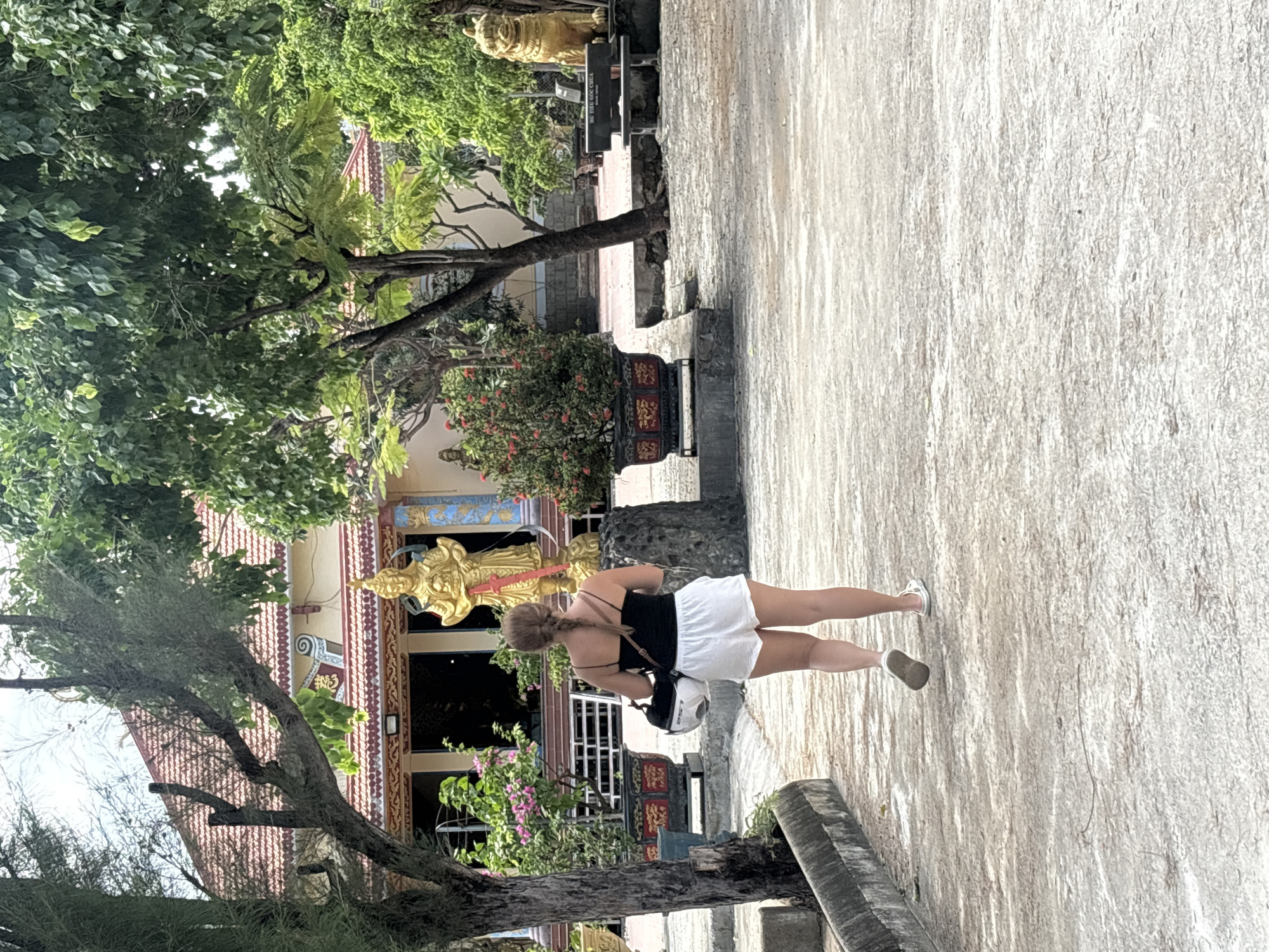 This image captures an outdoor scene at what appears to be a temple or cultural site, located in Cam Ranh, Vietnam. A woman, seen from behind, is the primary subject, walking on a light-colored, textured paved path. She is dressed in a black sleeveless top and white shorts, with light-colored shoes. Her hair is styled in a braid, and she is holding a white helmet in her left hand while also carrying a dark crossbody bag.
In the background, a building with a red-tiled roof is prominently featured. Its facade is richly decorated with intricate patterns in red, gold, and blue, and features several golden statues or relief sculptures, including a large, ornate figure near what appears to be an entrance. Dark windows or doorways are visible on the building's front.
The scene is framed by lush green trees with dense foliage, some of which appear tropical. Flowering bushes with pink blossoms add splashes of color near the building. On the paved path, a decorative black planter adorned with red and gold motifs is present. Further back, near another golden statue, a black bench can be seen with white text, which reads "BU TUC COC GIA." The overall setting suggests a daytime visit to a serene and culturally significant location.