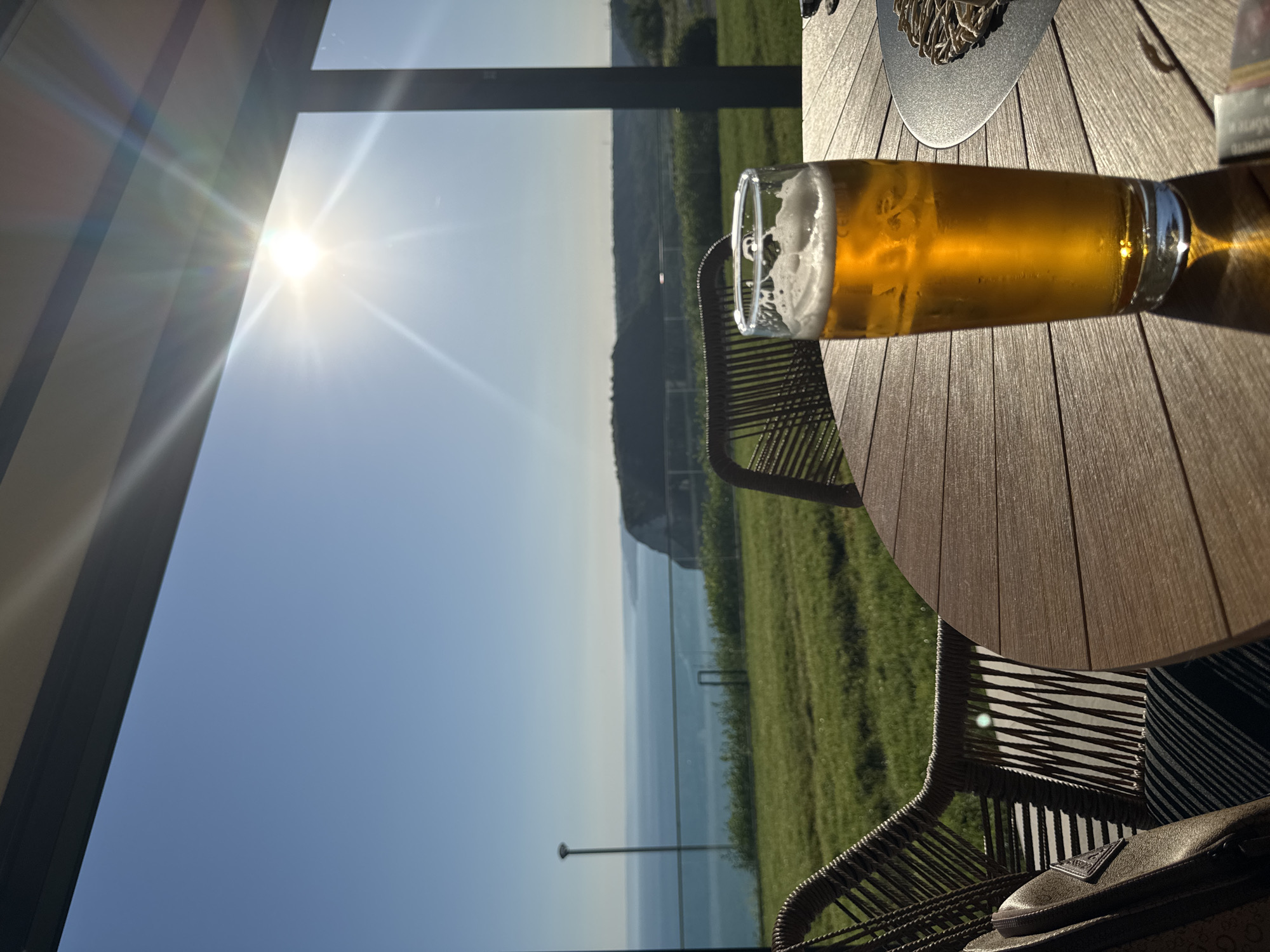 The image captures a serene scene outdoors, likely at a cafe or restaurant with a view. The primary subject is a glass of golden beer with a frothy head, positioned on a wooden table. The table surface has a distinct planked design. To the left of the table, a portion of a woven chair is visible, and a dark bag with a triangular logo rests on it.
The background reveals a breathtaking vista: a clear blue sky transitions into a hazy horizon overlooking a body of water, possibly the sea. Beyond the water, rolling green hills are visible, suggesting a rural or coastal setting. The sun is bright and high in the sky, casting lens flares and strong shadows, indicating it is daytime, likely late morning or early afternoon. The weather appears to be sunny and pleasant.
The scene evokes a sense of relaxation and enjoyment, perhaps someone taking a break to savor a drink while admiring the view. The focus on the beer and the scenic backdrop suggests a leisurely moment. The location context indicates this might be Kavarna, Bulgaria, known for its coastal beauty. There is no visible text in the image.
