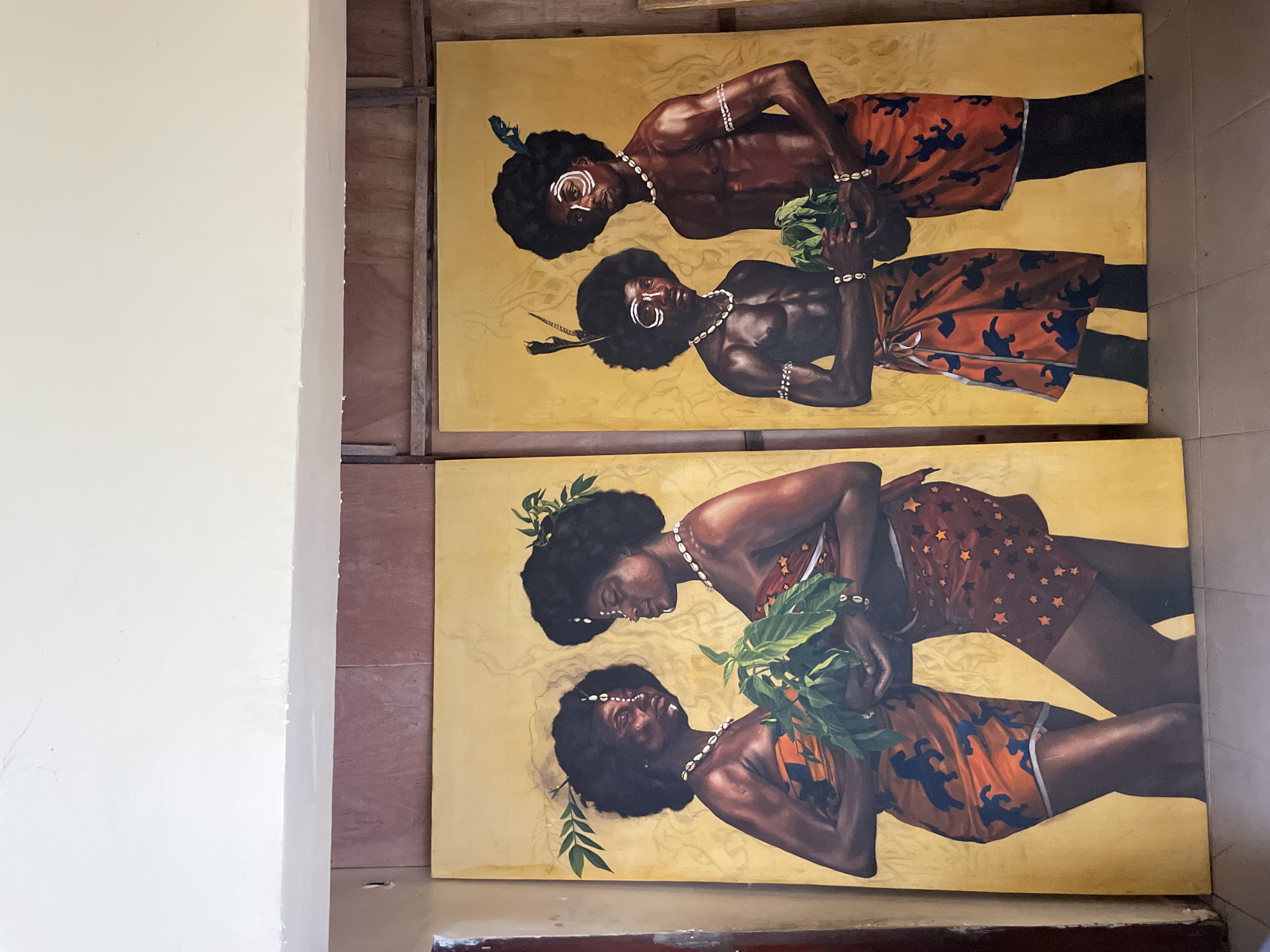 The image shows two large rectangular paintings leaning against a wall in an indoor space. The paintings are stacked vertically. Both feature two depictions of dark-skinned individuals in tribal attire. The background of each painting is a golden hue. The individuals in the top painting wear tribal face paint, shell necklaces, and armbands, and hold green foliage. They wear patterned orange and blue skirts. The bottom painting depicts individuals with leaves in their hair and shell necklaces, also holding foliage. Their skirts are patterned, one with stars and the other with animal silhouettes. The location appears to be an art studio or gallery setting in Ado, Nigeria.