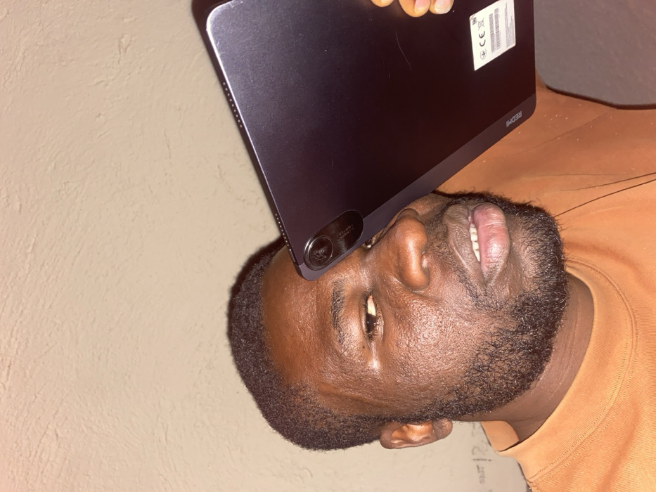 In the city of Udu, Nigeria, an adult male with dark skin, short dark hair, and a neatly trimmed beard is depicted. He wears an orange long-sleeved shirt. A dark gray Xiaomi Redmi tablet is held diagonally in front of his face, obscuring his left eye and upper forehead. The tablet's multi-lens rear camera module is visible near his right temple. The word "Redmi" and a white sticker with regulatory markings are discernable on the tablet's back surface. The man's right eye is partially visible, and his mouth is slightly open, revealing his lower teeth. The background consists of a plain, light-colored textured wall, illuminated by artificial light.