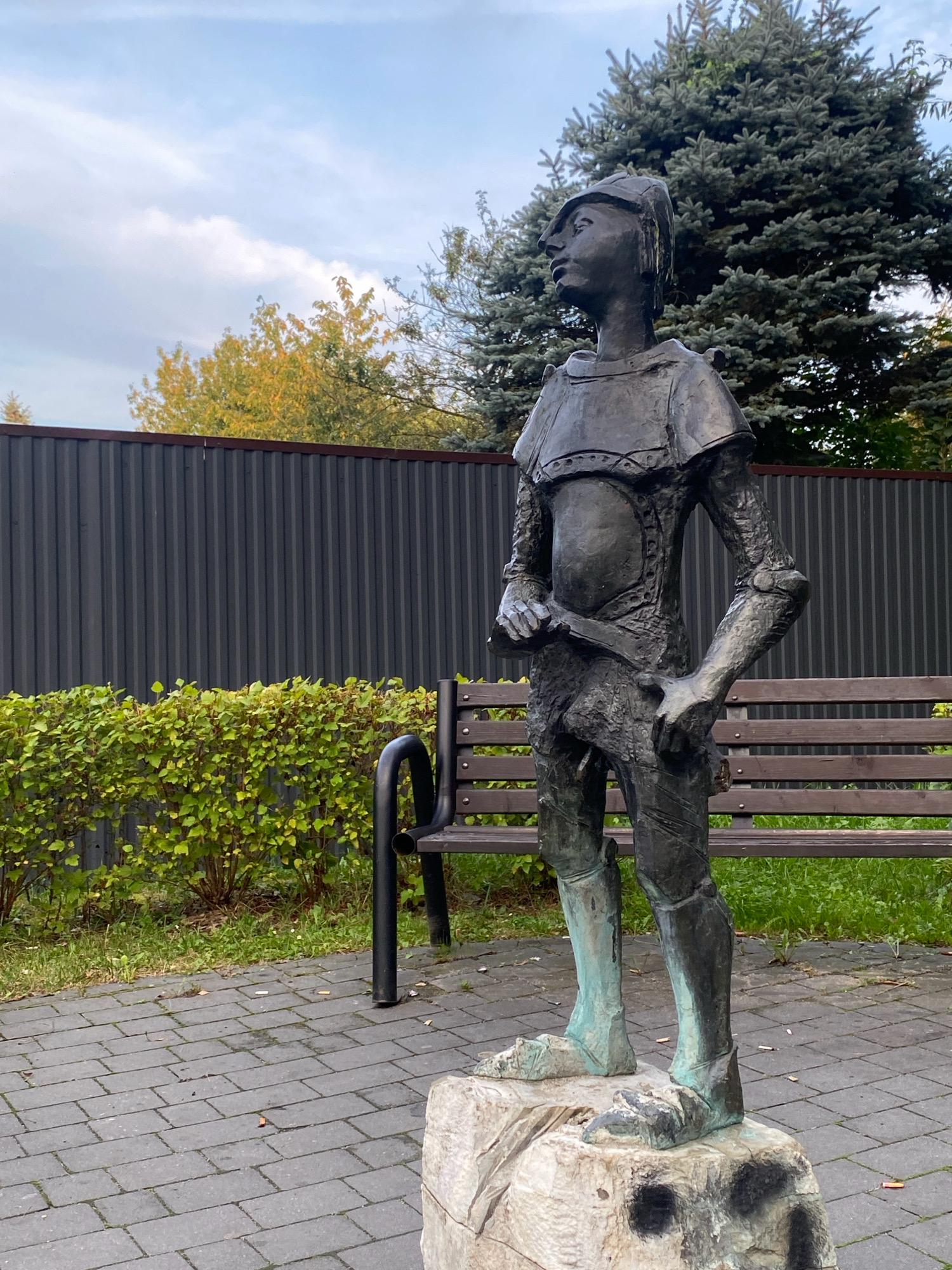A full-body bronze statue of a young person in what appears to be armor stands on a rough, light-colored stone base in front of a dark, corrugated metal fence. The statue is facing to the left in profile, looking upwards and to the left. It wears a helmet, a segmented chest plate, arm guards, and leg coverings. The statue's right hand is near its hip, and its left hand is holding a sword or similar object at its waist. The statue's lower legs and feet have a greenish patina. Behind the statue, a wooden bench with dark metal legs is visible, situated behind a dense, green hedge. Beyond the hedge, the dark metal fence stretches across the background. To the left of the statue, deciduous trees with yellowing leaves are visible, and to the right, a large evergreen tree stands. The ground in front of the bench and statue is paved with grey rectangular bricks, with scattered green grass and some cigarette butts. The sky above is partly cloudy with a mix of blue sky and white clouds.
The statue is located in Nowy Sącz, Poland, near the Municipal Sports and Recreation Center (MOSiR) on ul. Nadbrzeżna 76. It is a representation of the character of "Chłopiec z gwizdkiem" (The Boy with the Whistle) from the novel "Przygody Tomka Sawyera" (The Adventures of Tom Sawyer) by Mark Twain, depicted as a street urchin with a sword.