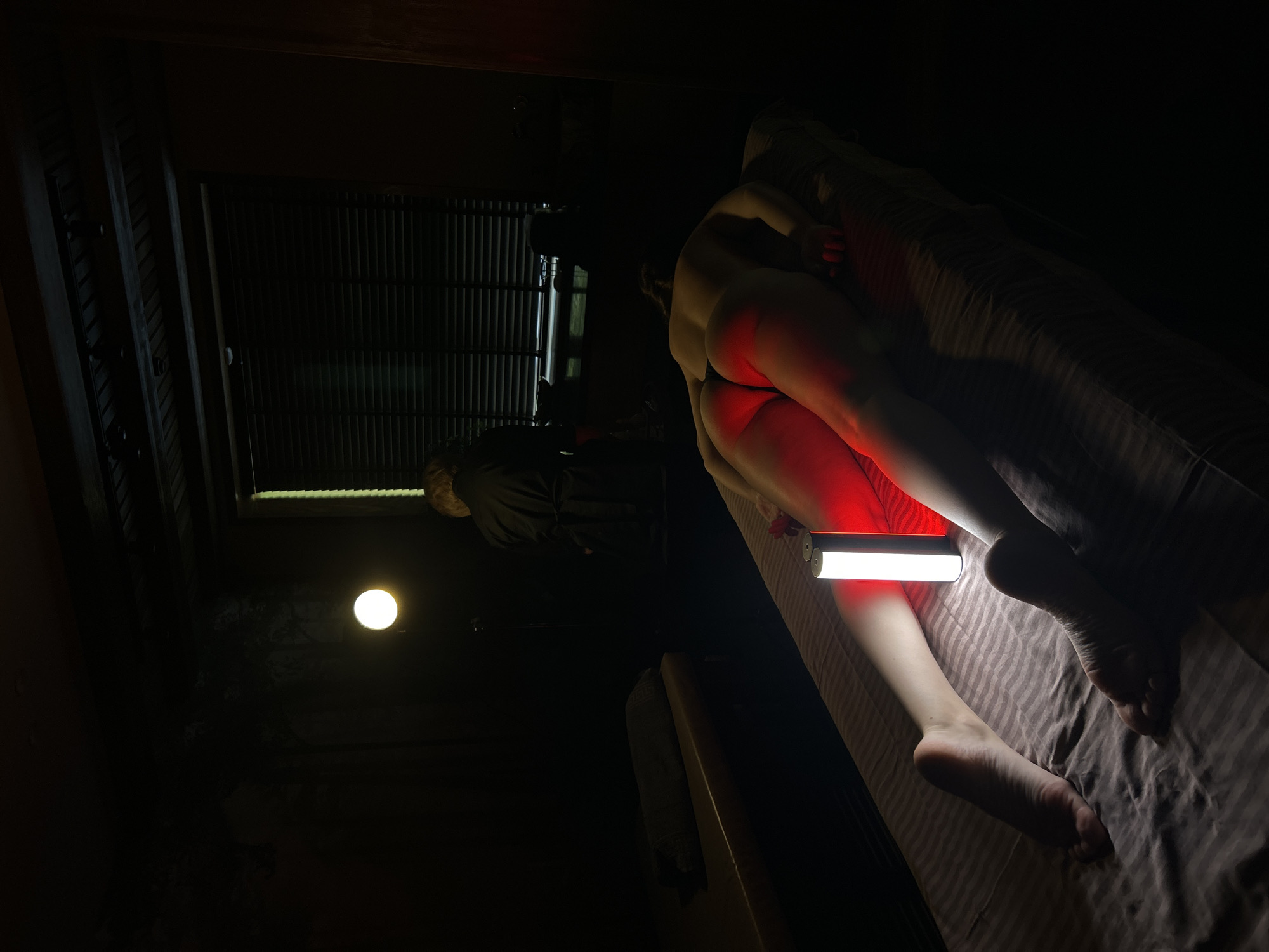 A dimly lit indoor scene shows a nude person lying on a bed under a red light. The person is positioned lengthwise on a bed covered in striped bedding. A small, rectangular red light shines on their lower legs. In the background, a window with blinds is visible. A light fixture illuminates the room from the upper left corner. Another person, wearing dark clothing, is standing further back in the room. There are no indications that this scene takes place in Izhevsk, Russia.