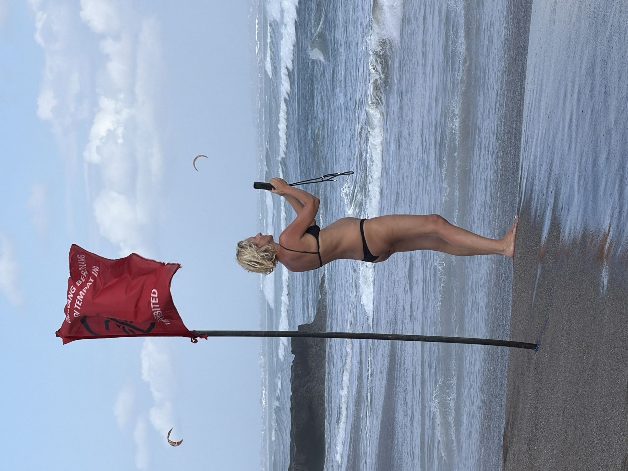 A Caucasian woman with blonde hair, wearing a black bikini, stands on a sandy beach, holding a mobile phone with a lanyard, taking a picture. To her left, a tall, thin, dark pole is planted in the sand. A large red flag, with white text and a symbol, is attached to the pole and is fluttering in the wind. The beach overlooks a choppy ocean with waves and some rock formations in the distance. Two kite surfers are visible in the sky. The location is identified as North Kuta, Indonesia, likely on a beach with rough water conditions as indicated by the red flag. The sky is partly cloudy.