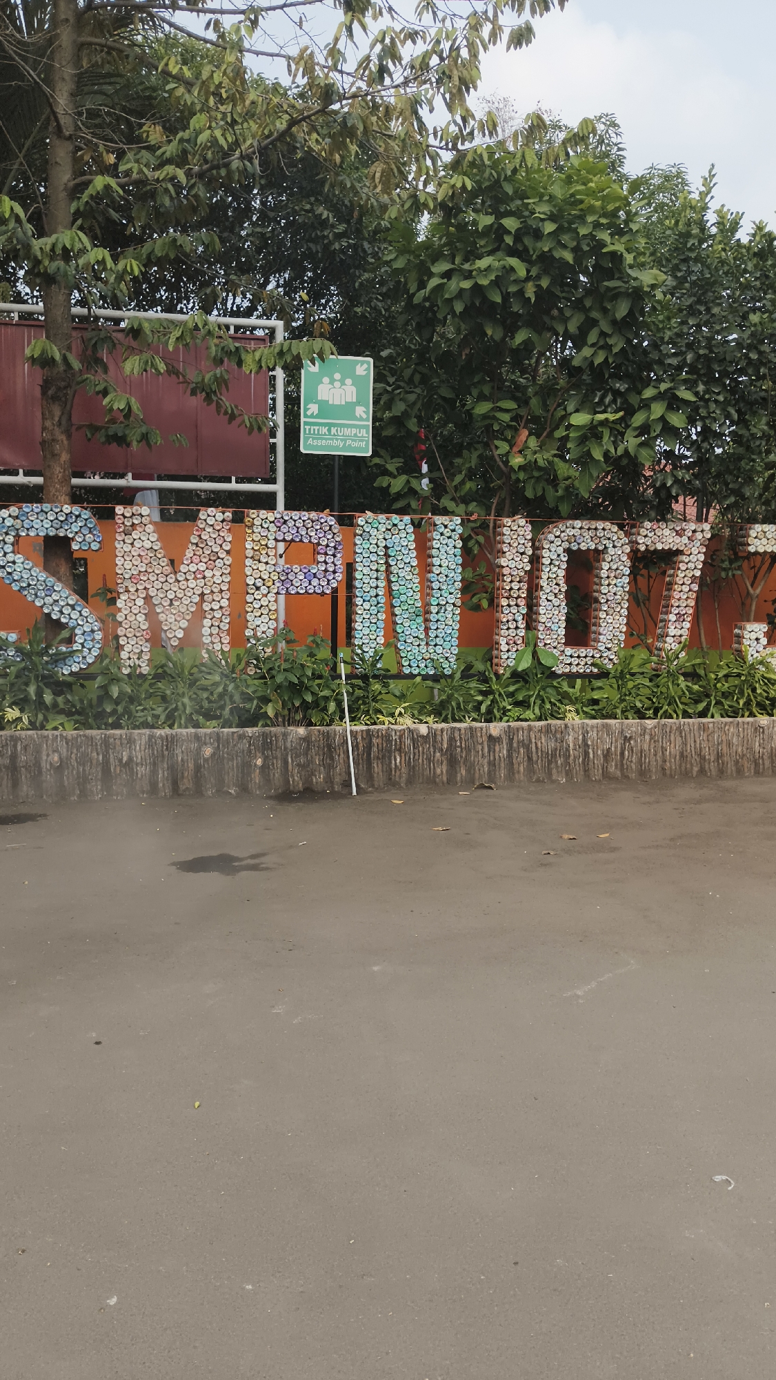 The image shows the exterior of SMP Negeri 107 Jakarta, located in South Jakarta, Indonesia. The school's name, "SMPN 107," is displayed in large, colorful, three-dimensional letters. These letters are constructed from numerous bottle caps, giving them a mosaic-like appearance. Behind the letters, an orange facade is visible, and further back, trees and foliage create a natural backdrop. To the left of the school's name, a burgundy wall or building structure is partially visible. A green sign with white icons and text reading "TITIK KUMPUL Assembly Point" is mounted on a pole, indicating a designated meeting spot. The school grounds appear to be fronted by a paved road or asphalt area, with some fallen leaves and a white cane lying on the ground in front of the lettering. The overall scene suggests an outdoor environment on a cloudy day, with the school building and its signage as the main focus.