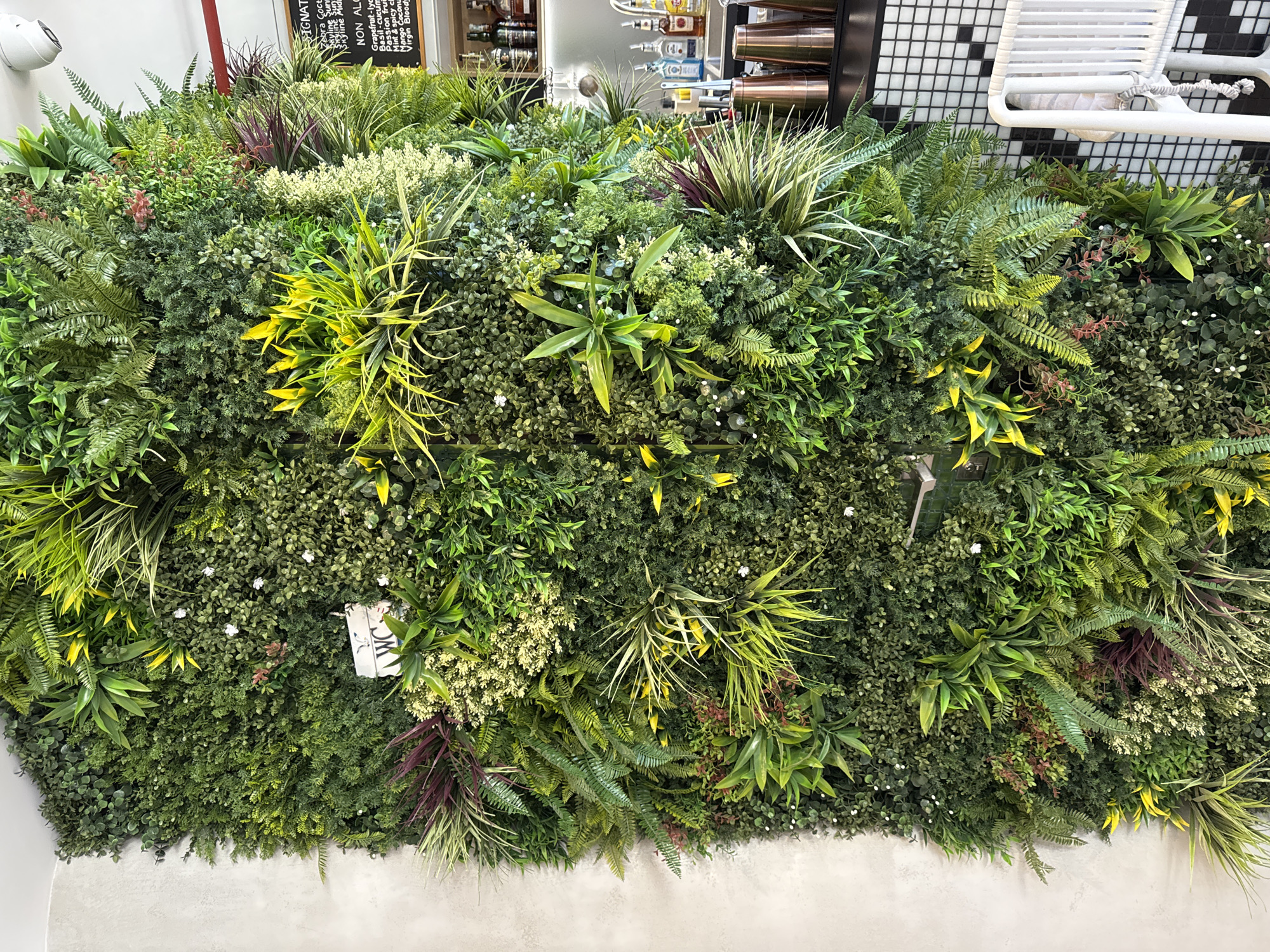 The image depicts a dense artificial green wall, featuring a variety of simulated plant species, textures, and shades of green. The wall is not uniform, incorporating accents of yellow, purple, and small white flowering details to mimic natural variation. A "WC" sign is partially visible, seemingly integrated into the foliage.
In the background, a bar area is subtly visible. Shelves stocked with bottles and glassware are noticeable above the green wall. A black and white tiled wall pattern and a white rack structure, possibly a towel rack, are also present.
There is no indication of people. The focus is on the constructed green environment. The image originates from an indoor location with bar elements, likely in an establishment with a deliberate nature-inspired or biophilic design. No clear evidence indicates this is in Boreti, Montenegro.