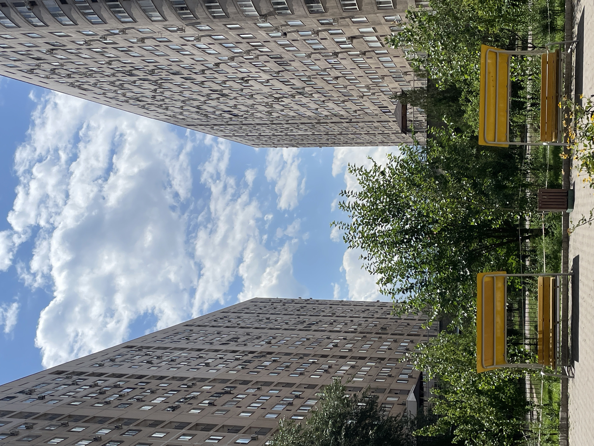 The image depicts a view between two identical, tall, beige apartment buildings in Almaty, Kazakhstan, on a sunny day with white, puffy clouds in a blue sky. In the foreground, two bright yellow metal park benches are positioned symmetrically, facing each other. Lush green trees and foliage are abundant in the middle ground, partially obscuring a small, dark gazebo structure visible behind the benches and between the apartment buildings. The ground is paved with light gray square tiles. The architectural style of the buildings is characteristic of Soviet-era mass housing, commonly found in Almaty. No people are present in the frame.