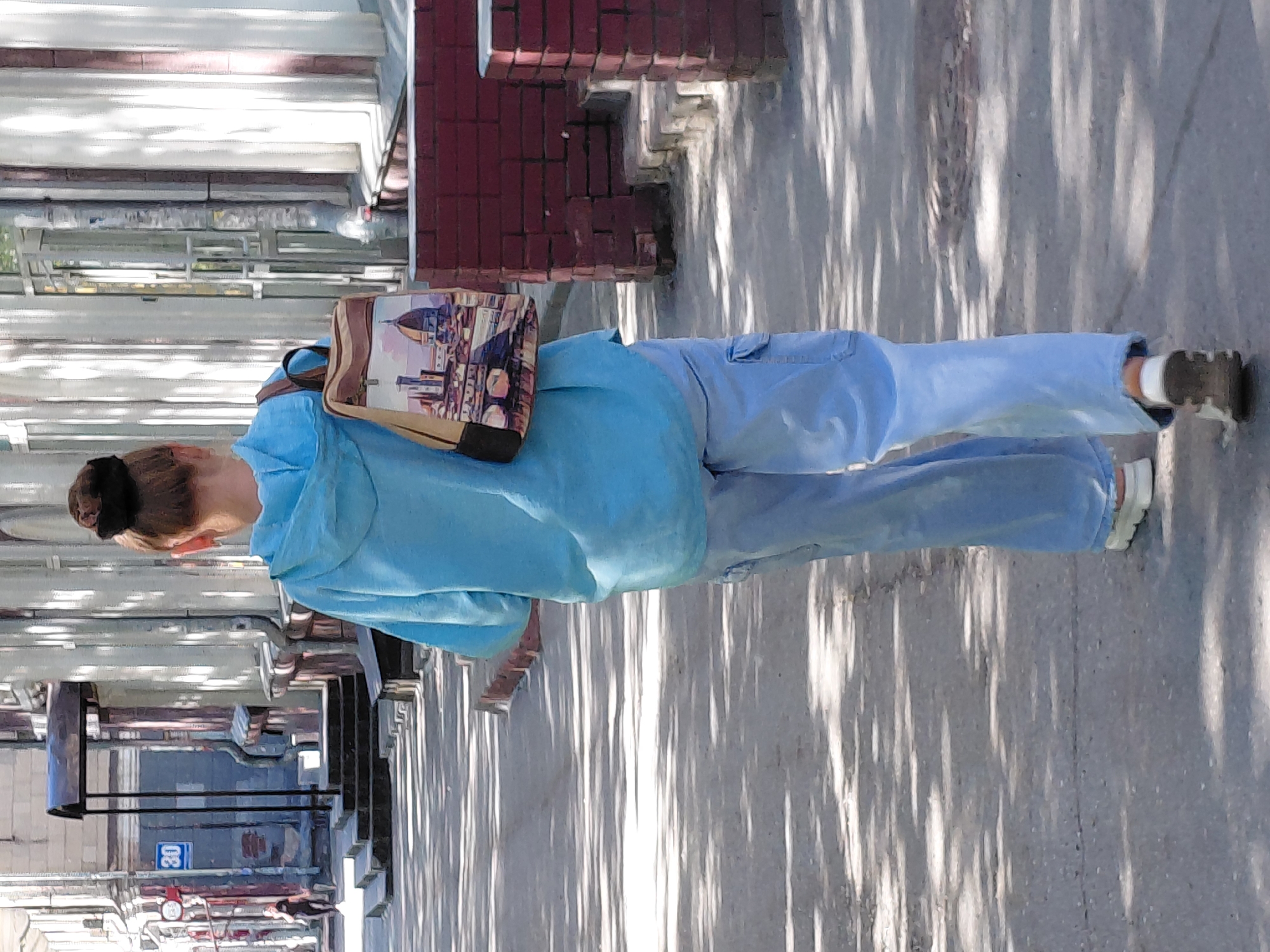 A person walks away from the camera on a sidewalk in an urban environment. The person is wearing a light blue hooded sweatshirt and light blue cargo pants. They have a backpack on their shoulders with an image of a cityscape on it. The sidewalk is made of asphalt and is lined with shadows from trees. To the right of the sidewalk is a low red brick wall. To the left of the sidewalk are the facades of buildings with columns and windows. In the background, there is a street sign with the number 30 on it, and a person walking in the distance. The lighting suggests it is daytime with sunlight filtering through trees.