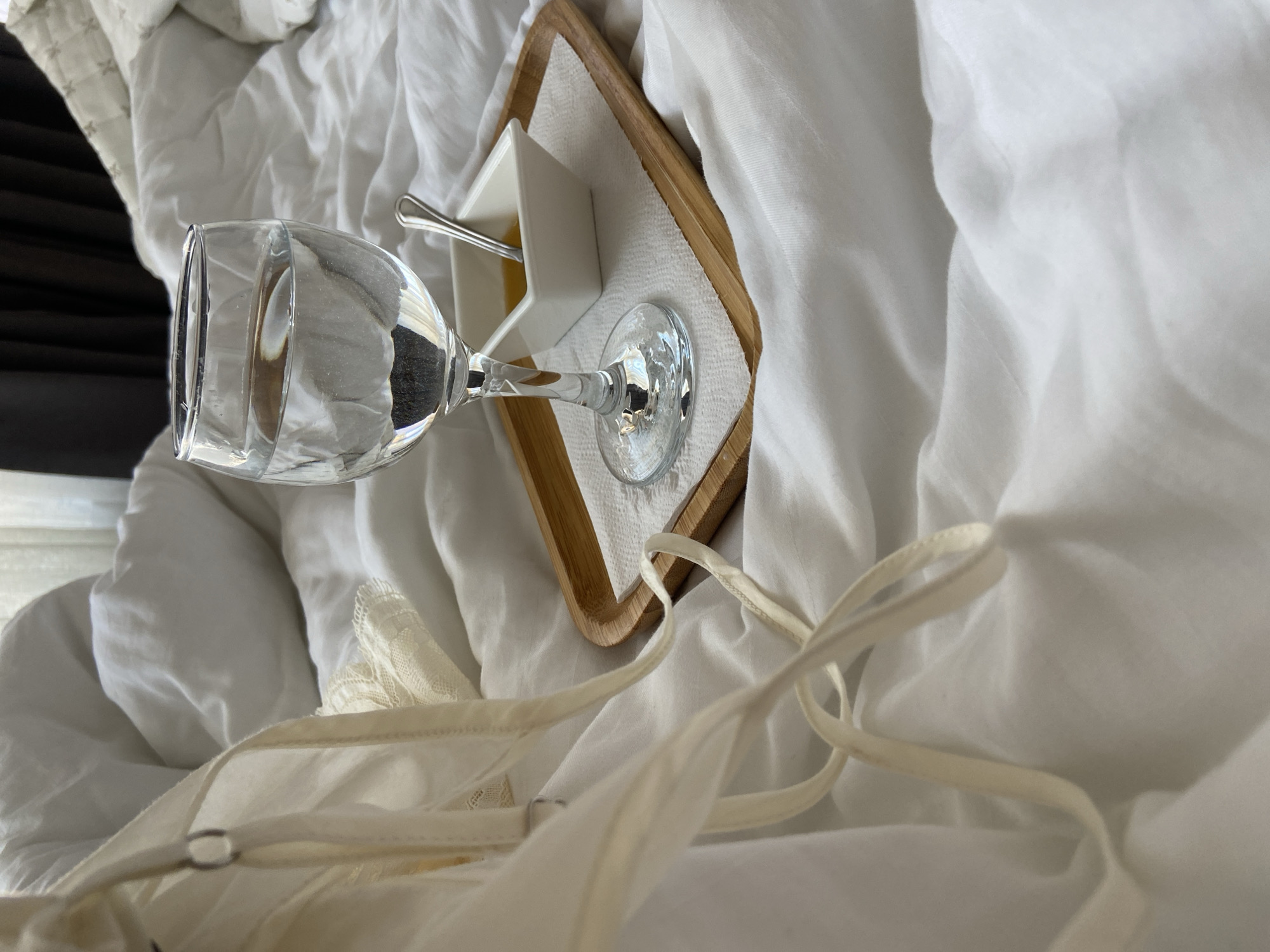 The scene depicts an indoor environment in Budva, Montenegro, focusing on a rumpled bed covered with white bedding. Centered on the bedding is a light wooden rectangular tray. On the tray, a clear stemmed glass filled with water stands on a white paper napkin. Adjacent to the glass, also on the napkin, is a small white square ceramic dish containing a yellowish liquid, consistent with honey, and a small silver spoon. To the left foreground, light-colored fabric straps, possibly from clothing or sleepwear, are draped across the white bedding, with a small silver ring visible on one strap. A section of light-colored lace fabric is also visible on the bedding near the straps. In the upper right background, dark curtains are partially visible, indicating a window beyond. No individuals are present in the image.