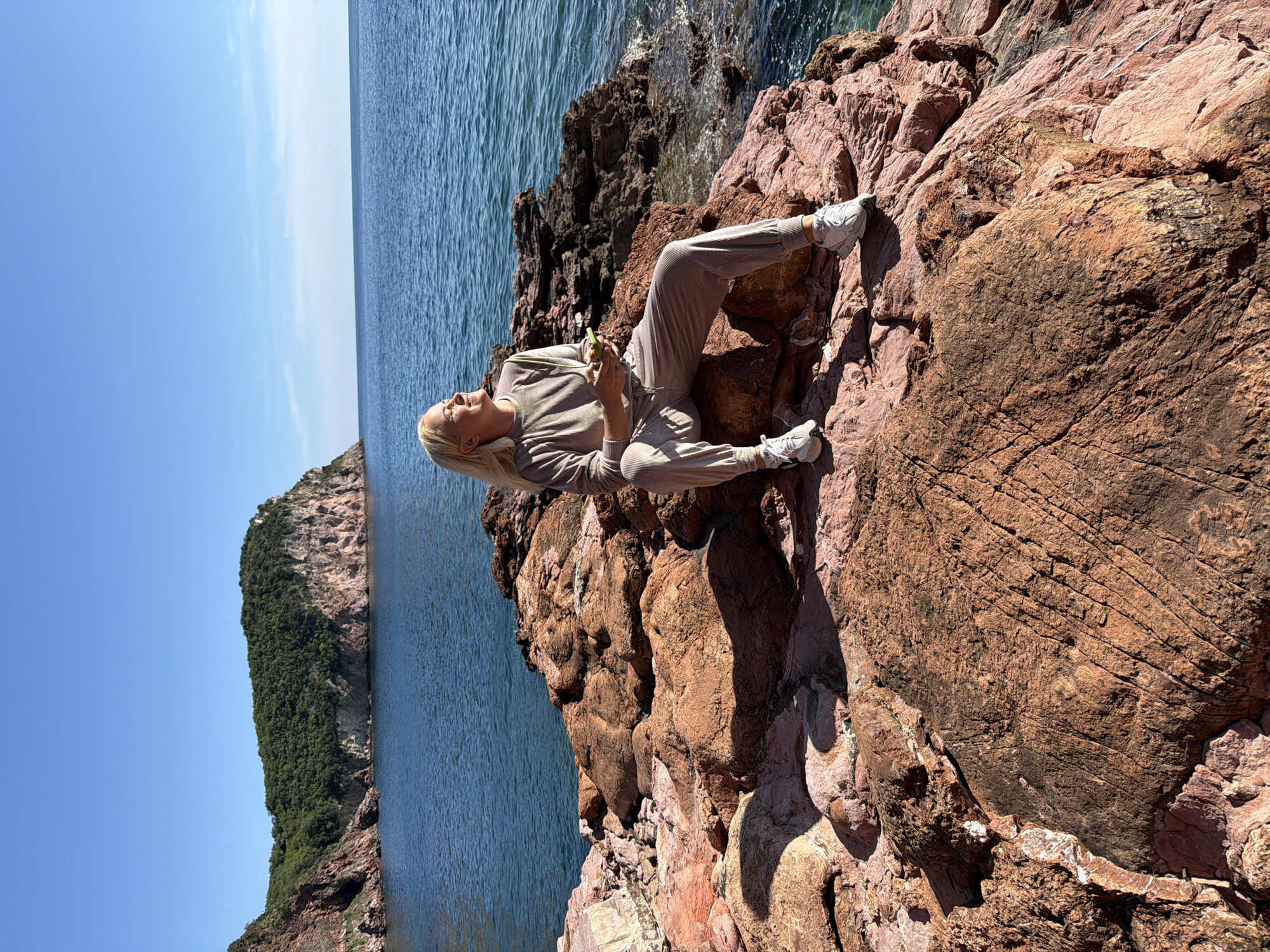 A woman is reclining on a rocky outcrop, potentially part of a cliff, situated near the coastline of Sveti Stefan, Montenegro. She is lying diagonally across the frame, dressed in a matching taupe-colored sweat suit and white sneakers. Her blonde hair is loose, and she appears to be looking up and away. In her hands, she holds a small, green object, possibly a piece of fruit or a small container.
The rocks surrounding her are reddish-brown and appear weathered. Beyond the rocks, the sea is visible, a deep blue color, with a small island or peninsula covered in green vegetation and reddish rock formations jutting out of the water in the distance. The sky above is clear blue. The scene is well-lit, suggesting a sunny day.