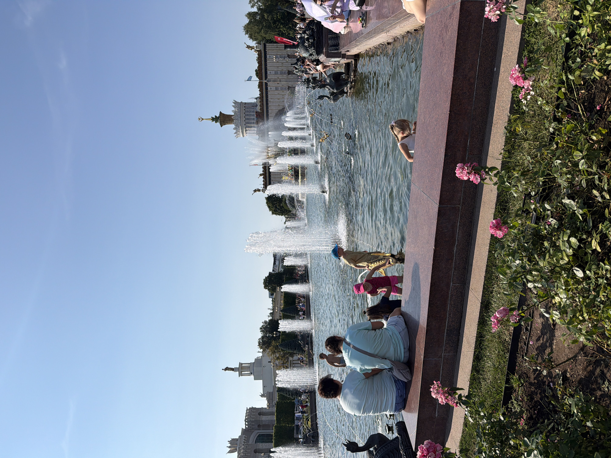 The image depicts the Friendship of Nations Fountain at VDNKh in Moscow, Russia. The fountain features numerous water jets spraying into the air against a backdrop of grand Soviet-era architecture. In the foreground, several people are enjoying the water. Two adults are seated on the edge of the fountain's basin, facing away from the camera. In the water, a young child in a pink outfit and blue hat is playing. To the right of the child, a young boy in a brown shirt and blue hat is standing. A young girl in a white top is leaning into the water on the right side of the frame. The fountain itself is adorned with statues, including swan figures. The sky above is clear and blue. Pink roses are blooming in the foreground, adding a touch of color to the scene.