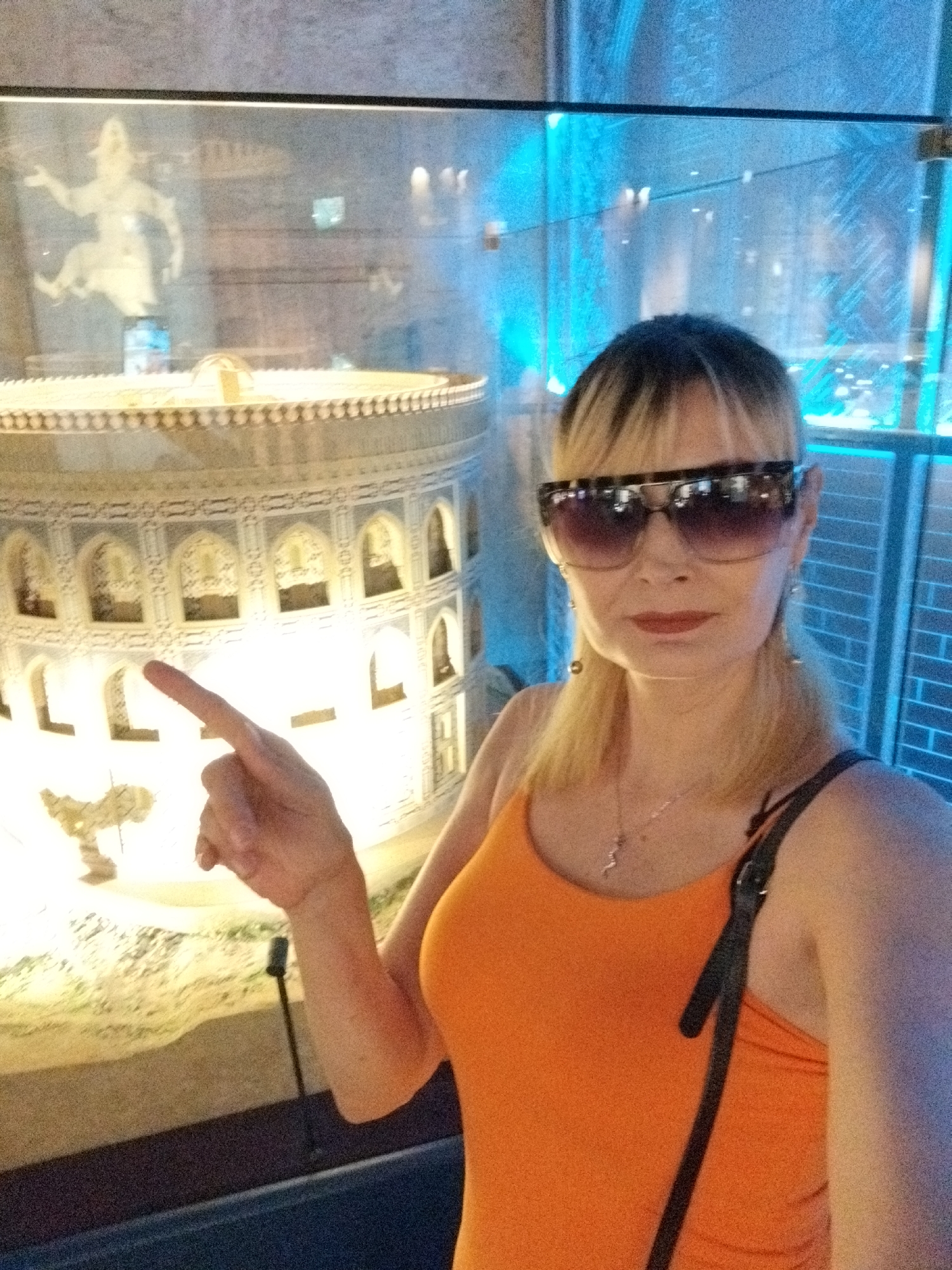 The image shows a selfie taken by a woman with blonde hair, wearing large sunglasses and an orange tank top, in front of a display case. Her right hand is extended, with her index finger pointing towards the display. Inside the display case is a miniature architectural model of a circular building with intricate carvings and arched openings. Behind the model, a pale statue of a figure in motion is visible, illuminated by blue lights. The background also shows the texture of a brick wall and a glass partition. The overall scene suggests an indoor exhibition or museum setting.