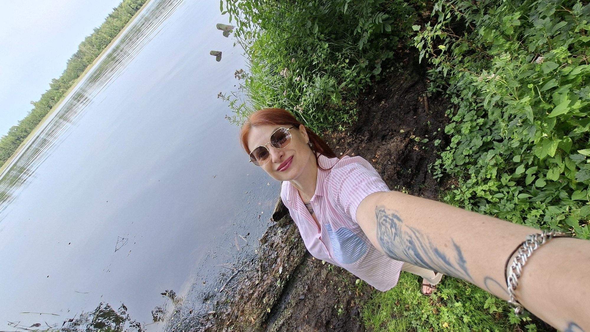 A woman with red hair and sunglasses is taking a selfie on the shore of a lake, with lush green vegetation on her right and the water stretching out before her. The trees in the background are dense and line the distant shore. The woman is wearing a pink and white striped button-down shirt and has a tattoo on her arm. A silver bracelet is also visible on her wrist. The location appears to be a natural setting, possibly a park or wilderness area, consistent with the description of Мядзельскі сельскі Савет, Belarus.