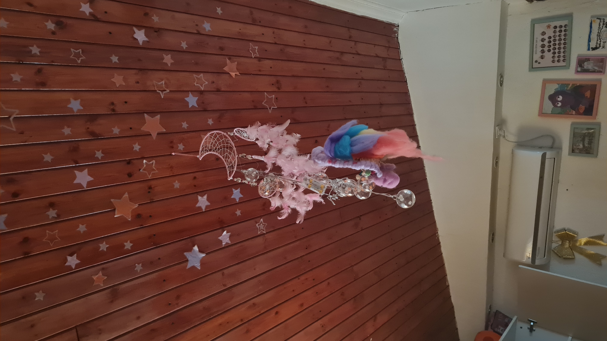 The image shows a room in Lachish Regional Council, Israel. The room features a brown wooden wall decorated with star-shaped decals. Suspended from the wall is a decorative object composed of a crescent-shaped dreamcatcher, pink feathers, rainbow-colored wool, and small glass orbs. The adjacent white wall is adorned with several framed pictures and a small white air conditioning unit. A shelf below the air conditioner holds a small golden bow.