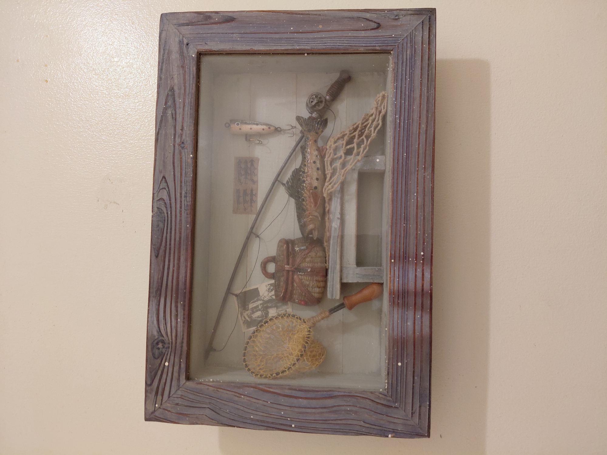 A rectangular shadow box with a rustic, painted wood frame hangs on a pale beige wall. The frame has a blue-grey wash with white speckles. Inside the box, arranged against a light-colored, possibly white or pale grey backdrop, is a collection of fishing-related items. These include a fishing rod, a fishing reel, a lure, a fish, and two nets. A small, woven creel and a photograph are also visible. The fishing rod is positioned diagonally, with the reel towards the top of the box. The fish stands upright with one net behind it, and one below. The presentation of these objects suggests a decorative or display purpose.
There is no information within the picture to suggest this image was taken in Cutler Bay, United States of America. This location has been given by request.