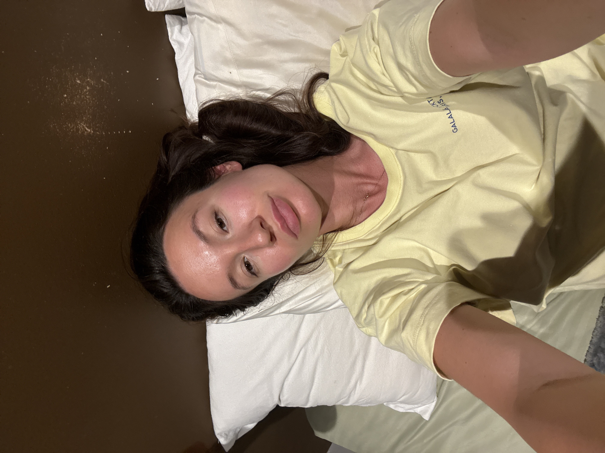 A woman lies on a bed with a white pillow and light green sheets. She is wearing a light yellow t-shirt with blue lettering that reads "GALALARIS". Her dark hair is partially pulled back. The background behind the bed is a dark brown wall with some speckles. The image appears to be a selfie taken by the woman. The setting is a bedroom. There is no information to suggest a specific location within Tel Aviv, Israel.