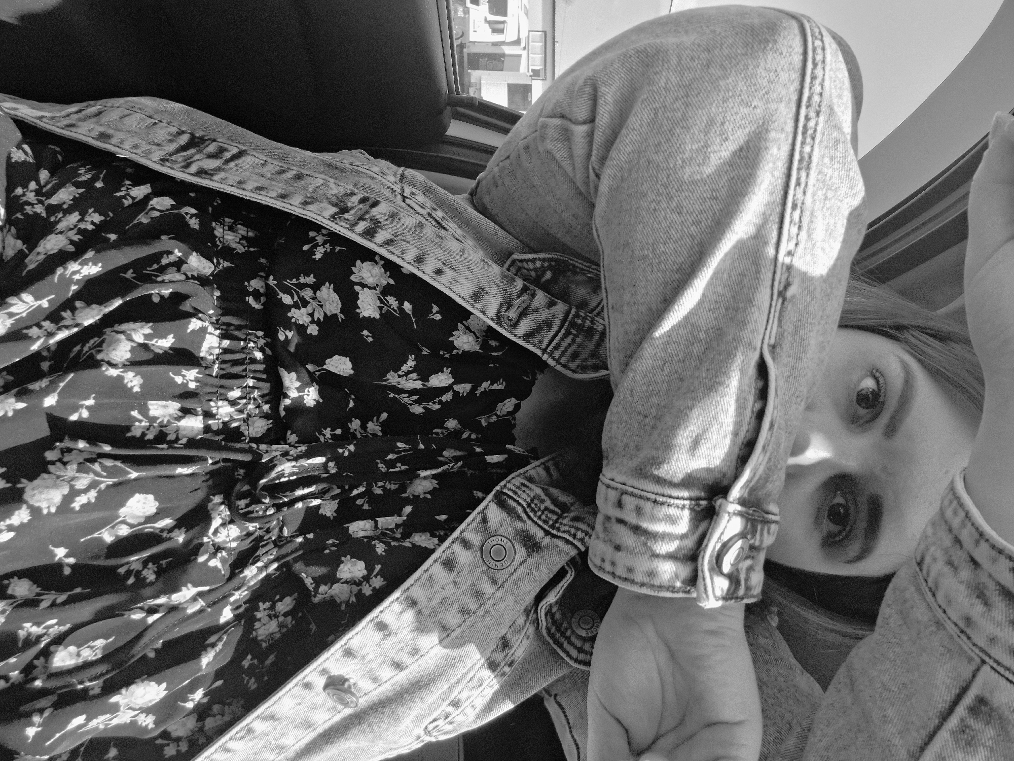 A monochrome, slightly distorted, high-angle shot looking down onto a person wearing a denim jacket over a floral print top. The jacket is unbuttoned, revealing the print of the top. The person's face is partially visible, showing their eyes looking up at the camera. Their hand is also visible in the foreground, near the cuff of the jacket. The setting appears to be indoors, possibly in a vehicle, as evidenced by the presence of a window and a glimpse of building exteriors in the background. Based on the information provided (Izhevsk, Russia), it can be inferred that the location is in Izhevsk, but no specific details in the image confirm a specific location.