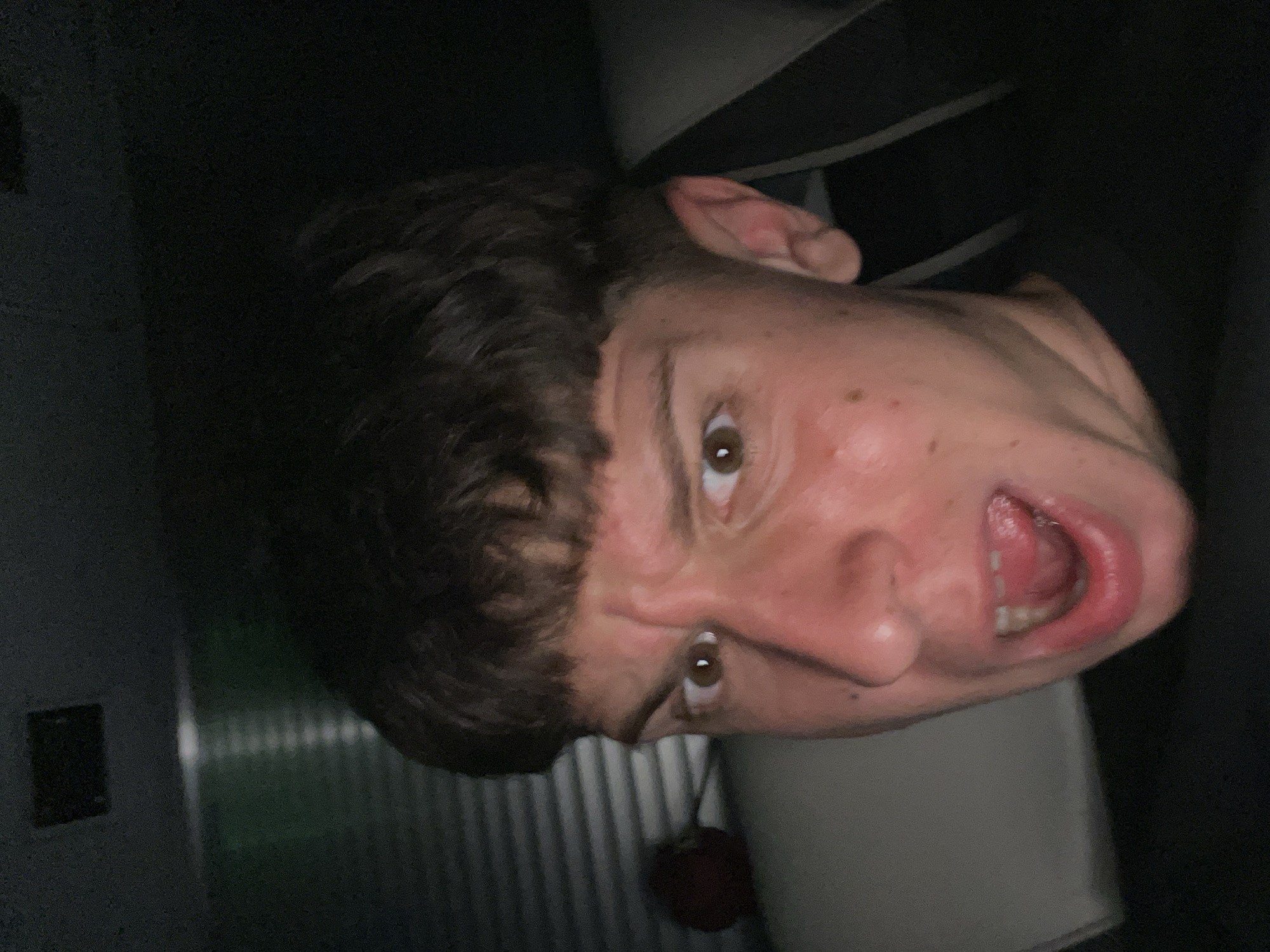 A close-up, distorted photograph shows a Caucasian male's face angled upward, mouth open with the tongue visible. He has short, dark brown hair with a fringe. His skin is light with visible pores and some red discoloration, likely from strain or emotion. Eyes are brown. Behind him are dark areas, with a vertically striped wall visible to the left and what appears to be a black and white striped garment. There is a red object hanging below his face. The location is not identifiable from the image.