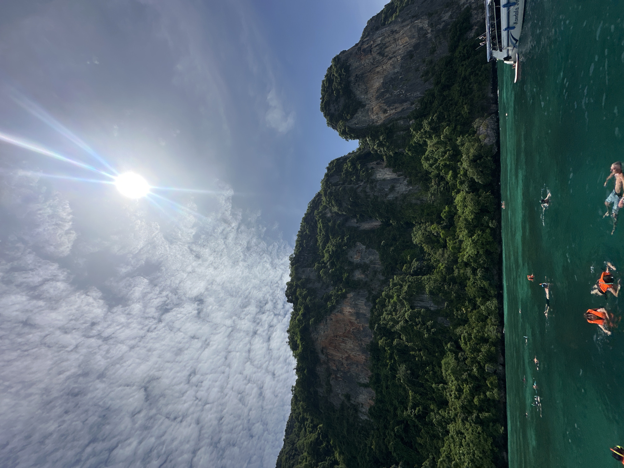 The image captures a scenic view of Ao Nang, Thailand, featuring a rocky, jungle-covered island rising from the clear, turquoise waters of the Andaman Sea. The sun shines brightly overhead, creating a lens flare and illuminating a sky dotted with wispy clouds. Several people are visible in the water, some snorkeling with visible masks and snorkels, while others are swimming or floating. An orange life jacket is worn by at least one individual. In the foreground, a portion of a white boat with blue trim and text that reads "Kandala Sea" is present, with a person lying on a surfboard attached to its side. The water is calm and transparent, allowing for a view of the seabed in shallower areas. The overall scene depicts a popular tourist destination, likely a snorkeling or boat tour.