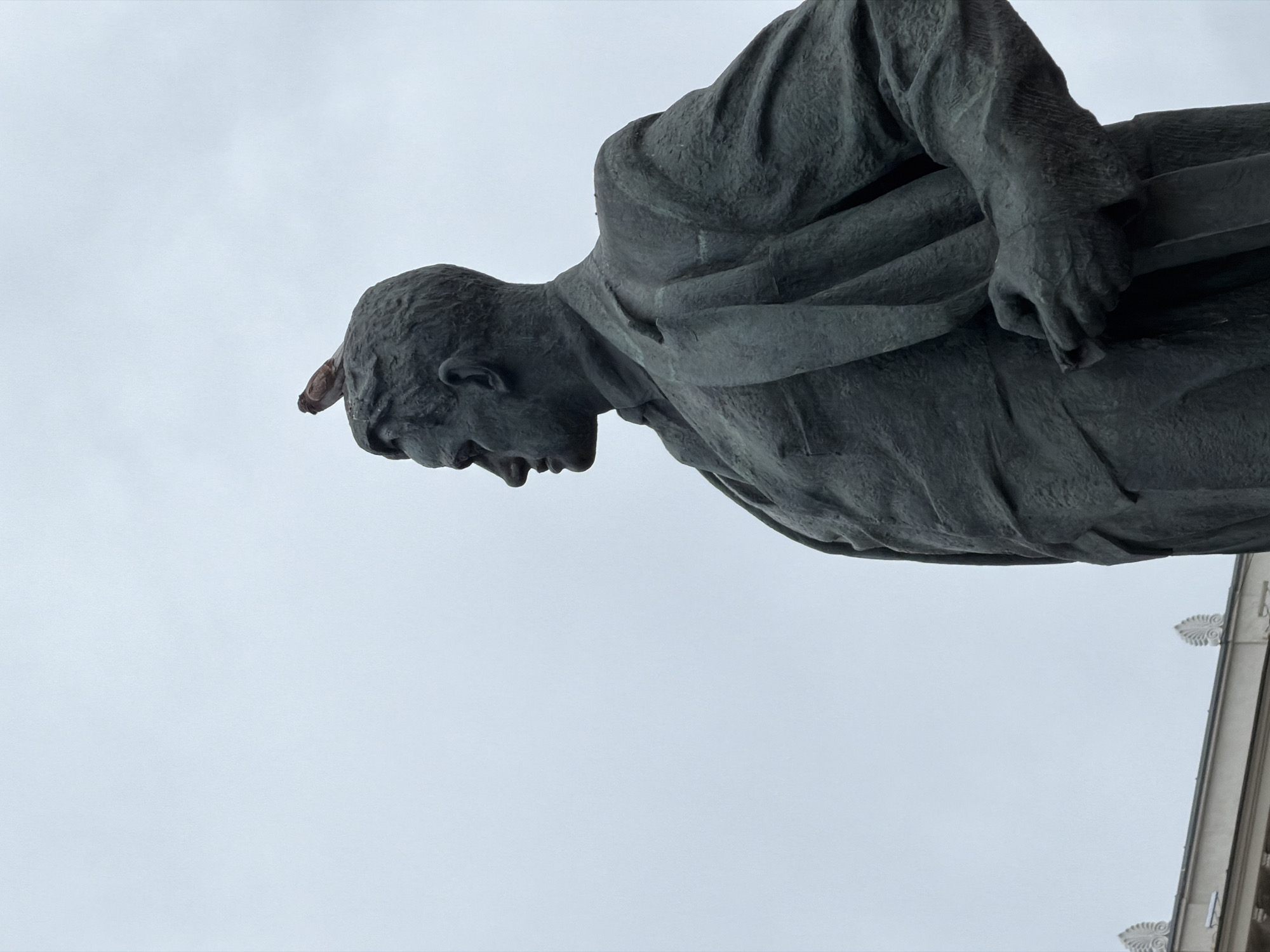 A low-angle shot of a weathered, dark grey statue of a man. The statue is positioned against a pale grey sky. A small, brown bird is perched on the head of the statue. In the background, the corner of a light-colored building with ornate, decorative elements is visible. This is a statue of Yuri Dolgoruky in Moscow, Russia.