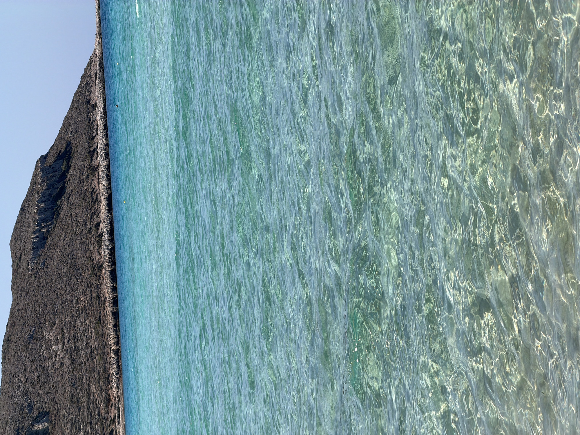 The image shows a view of the Aegean Sea in Greece, with clear turquoise waters and a brown, rocky hill in the background. The water is slightly rippled, and the seabed is visible in the shallower areas, appearing as a sandy or pebbly surface. The sky is clear and blue, suggesting a sunny day. There are no people or man-made objects clearly visible in the scene, other than possibly some small yellow buoys in the water. The image appears to be taken from a beach or shallow water area.