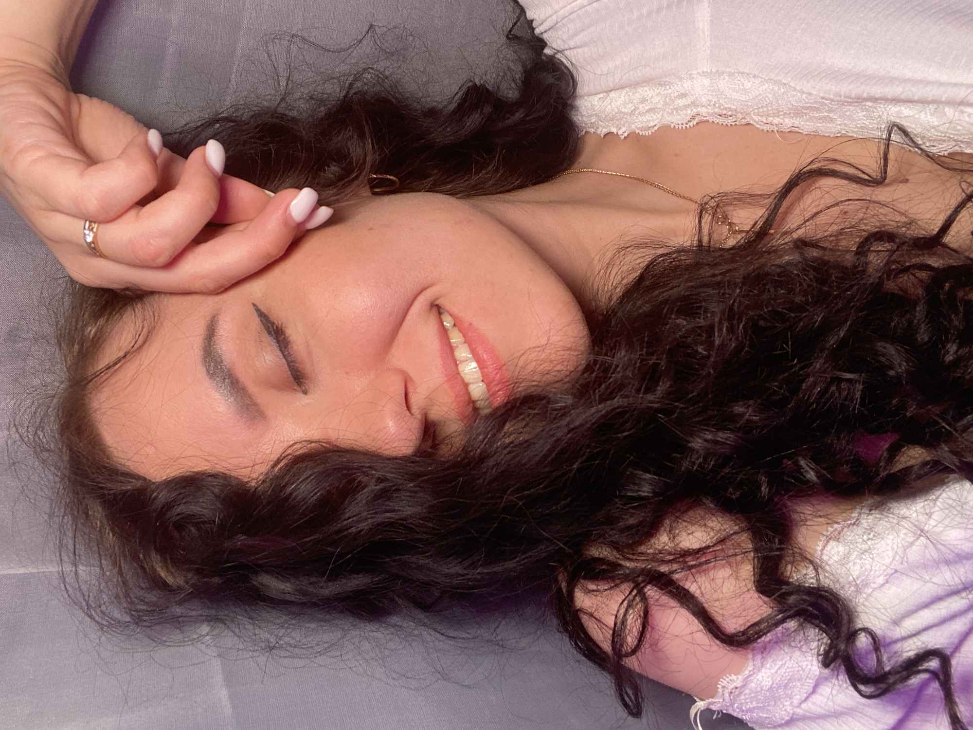 A woman is lying on a gray surface with dark brown curly hair spread around her face. She is wearing a white top with lace trim. Her right hand is raised near her face, with a ring visible on her ring finger and light-colored fingernails. Her eyes are closed, and she is smiling. The location is Chaikovsky, Russia.