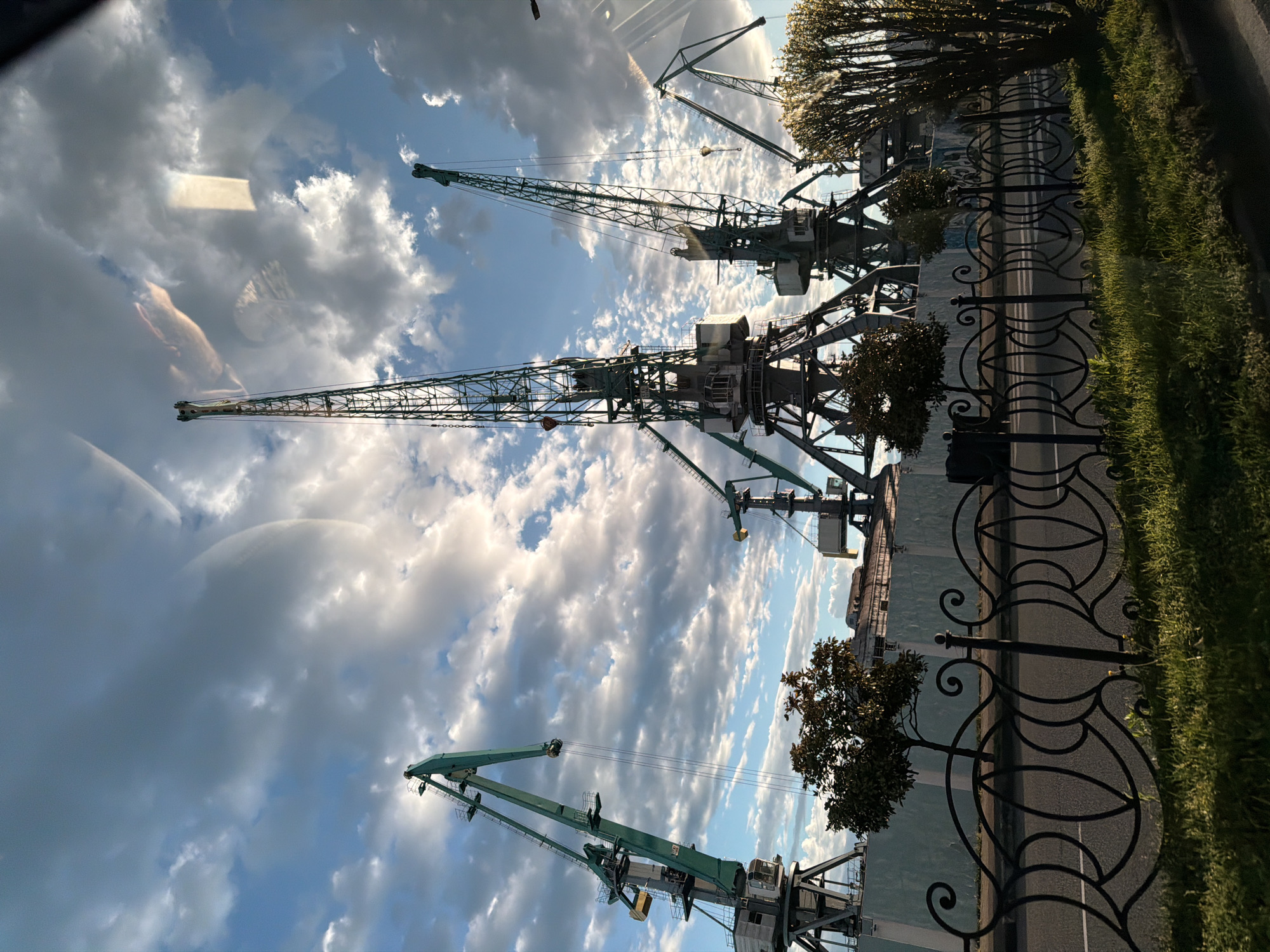 The image depicts a view of cranes in Batumi, Georgia, likely within or near the port area. Several large, green industrial cranes are visible against a backdrop of a cloudy sky. A decorative, wrought iron fence runs along the bottom right of the image, with small trees positioned behind it, bordering a road. Buildings are seen behind the cranes. There are some light reflections on the left side of the image.