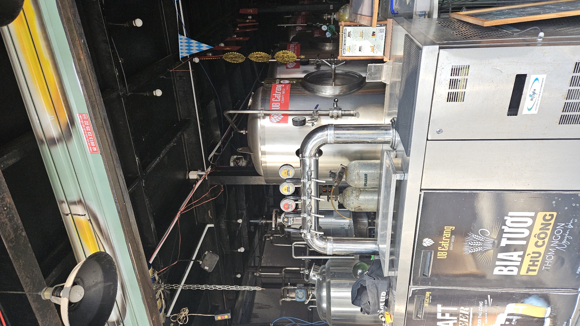 This image shows the interior of what appears to be a brewery or craft beer pub, likely located in Nam Nha Trang, Vietnam. Stainless steel brewing tanks and equipment are prominent, with associated valves, piping, and gauges. The brand "UB Eatrang" is displayed on the tanks and advertising panels, suggesting the name of the brewery or pub. Beer taps are visible. Labels on the tanks indicate the presence of "PRAXAIR" and "R134a," suggesting materials or equipment used in the brewing or cooling process. Advertising panels display text in Vietnamese, including "BIA TƯƠI THỦ CÔNG" and "THƠM NGÒN NGUYÊN BẢN," which translates to "fresh craft beer" and "fragrant, delicious, original." A shutter door with a phone number for repairs is visible. Overhead, a complex arrangement of wires, lights, and ceiling structures is visible. The scene conveys a sense of a working brewery or pub environment.