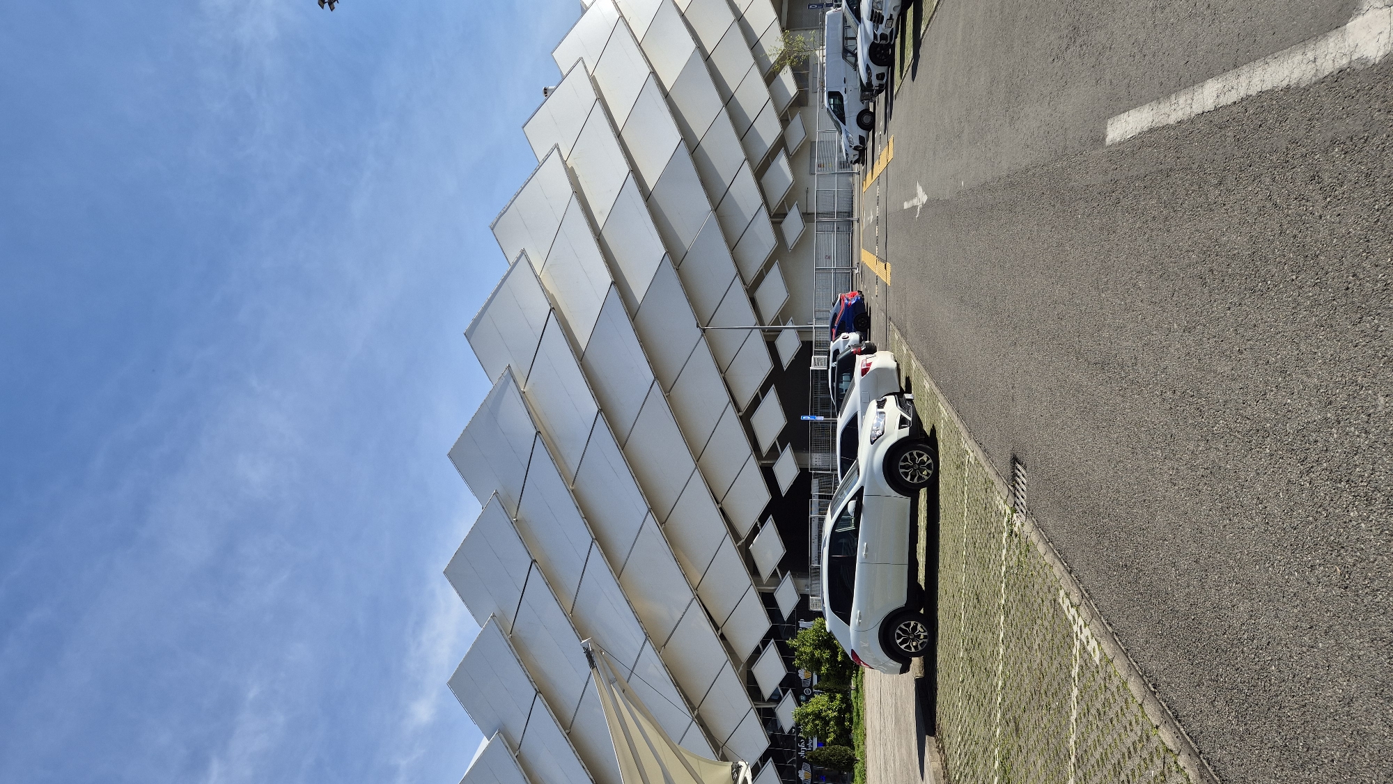 The image shows a section of the Piazza Batumi in Batumi, Georgia. The architectural facade is a multi-tiered diamond pattern design, constructed from what appears to be white or light-colored panels, ascending upwards and to the left. Adjacent to the building is a paved parking area with vehicles parked in a row parallel to the building. The sky is clear and blue with light cloud cover.