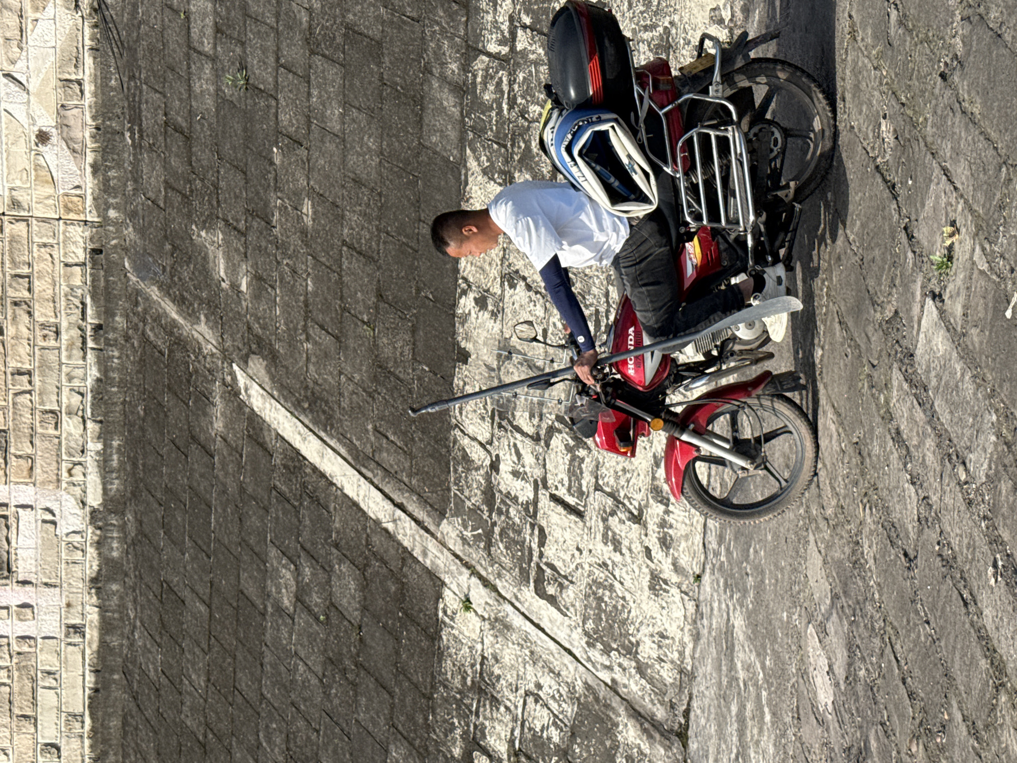 The image, rotated 90 degrees clockwise from its natural orientation, depicts a man and his red motorcycle parked on a broad, light-colored, patterned concrete or stone surface. To their right (if the image were oriented horizontally) is a substantial, dark grey, textured concrete or stone wall, likely an embankment or retaining wall, possibly along a river in Jiangjin, China.
The man, appearing to be of Asian descent, wears a white short-sleeved t-shirt over dark long-sleeved undershirts or arm sleeves, dark pants, and light-colored shoes. He has short, dark hair and is bending forward, holding a long, slender, light-colored pole or paddle with both hands. He appears focused on something in front of him, possibly adjusting the pole or preparing for an activity.
His motorcycle is a red "HONDA" branded vehicle, as indicated by text visible on its side. It features a black seat, a black top case or storage box mounted on a metal rack behind the seat, and another light blue and white bag (possibly a helmet bag or backpack with partial "POLICE" text) resting on the luggage rack. The motorcycle has chrome handlebars and front forks, and black wheels.
The setting suggests a sunny day, with clear light highlighting the textures of the concrete surfaces. The wall exhibits varying patterns, from rectangular blocks to more irregular, rougher sections, and some small weeds grow from its crevices. The overall scene conveys an outdoor, potentially riverside, environment in Jiangjin, China.
