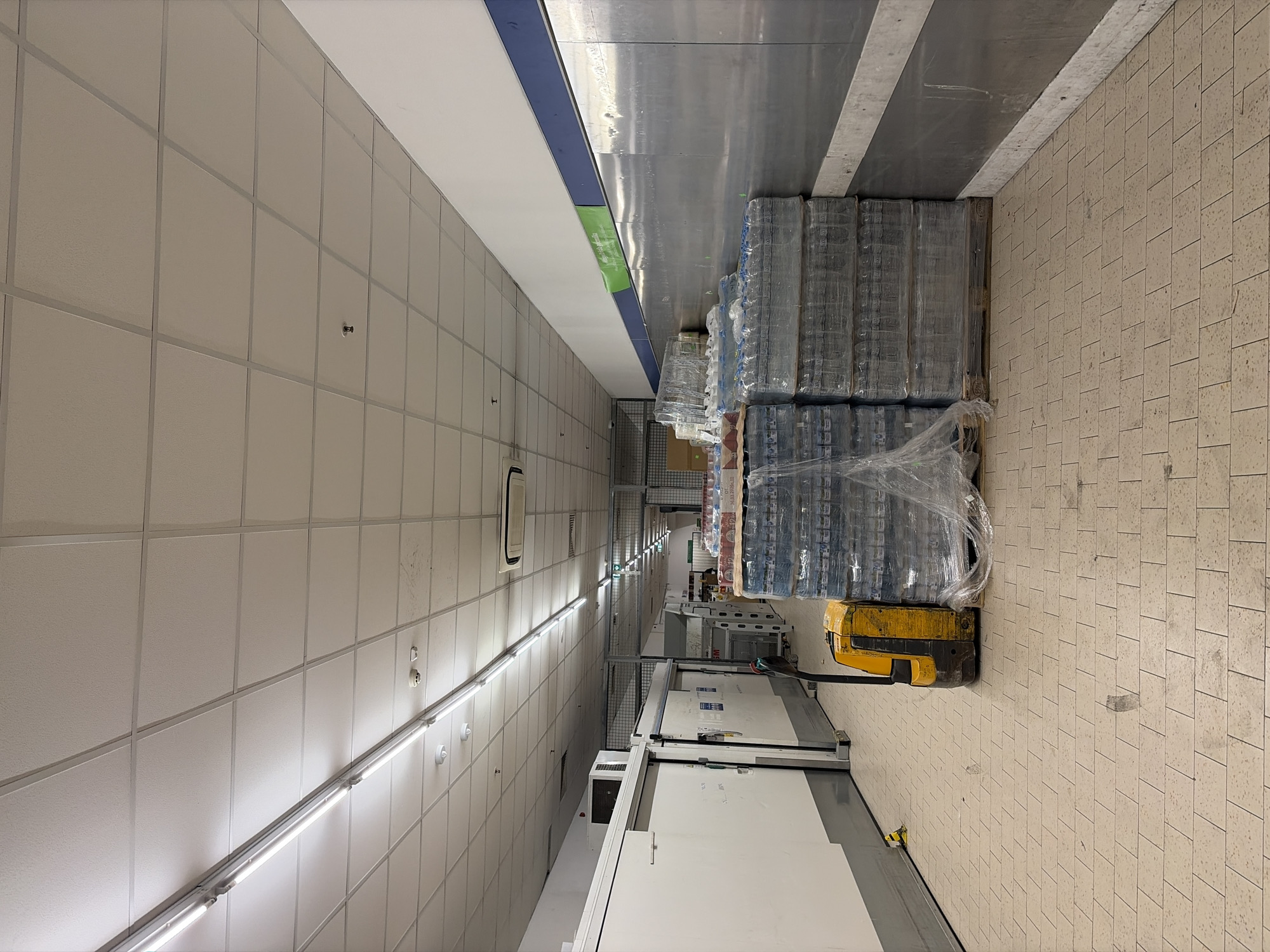 This interior image captures a storage or service corridor within a commercial establishment located in Plovdiv, Bulgaria. The floor is covered with light-toned rectangular tiles, exhibiting scuff marks and dirt. Along the left, a series of white modular panels, consistent with cold storage units or backroom facilities, feature prominent sliding doors and a mounted air conditioning unit. A yellow and black electric pallet jack is positioned on the floor, adjacent to the left-most stack of goods. Along the right, multiple pallets are stacked with shrink-wrapped clear plastic bottles of water, extending down the corridor. These pallets are positioned against a wall segment featuring a lower reflective metallic surface, a horizontal blue stripe, and an upper white section that displays a green sign with Cyrillic text, "Много ниска цена!" (Very low price!). The ceiling is a grid of white acoustic tiles, illuminated by numerous linear fluorescent light fixtures. Overhead, circular smoke detectors and a sprinkler head are visible, along with an air vent. The corridor recedes into the background, revealing additional industrial shelving, a metal mesh barrier, and a green exit sign. No individuals or active processes are present in the scene.