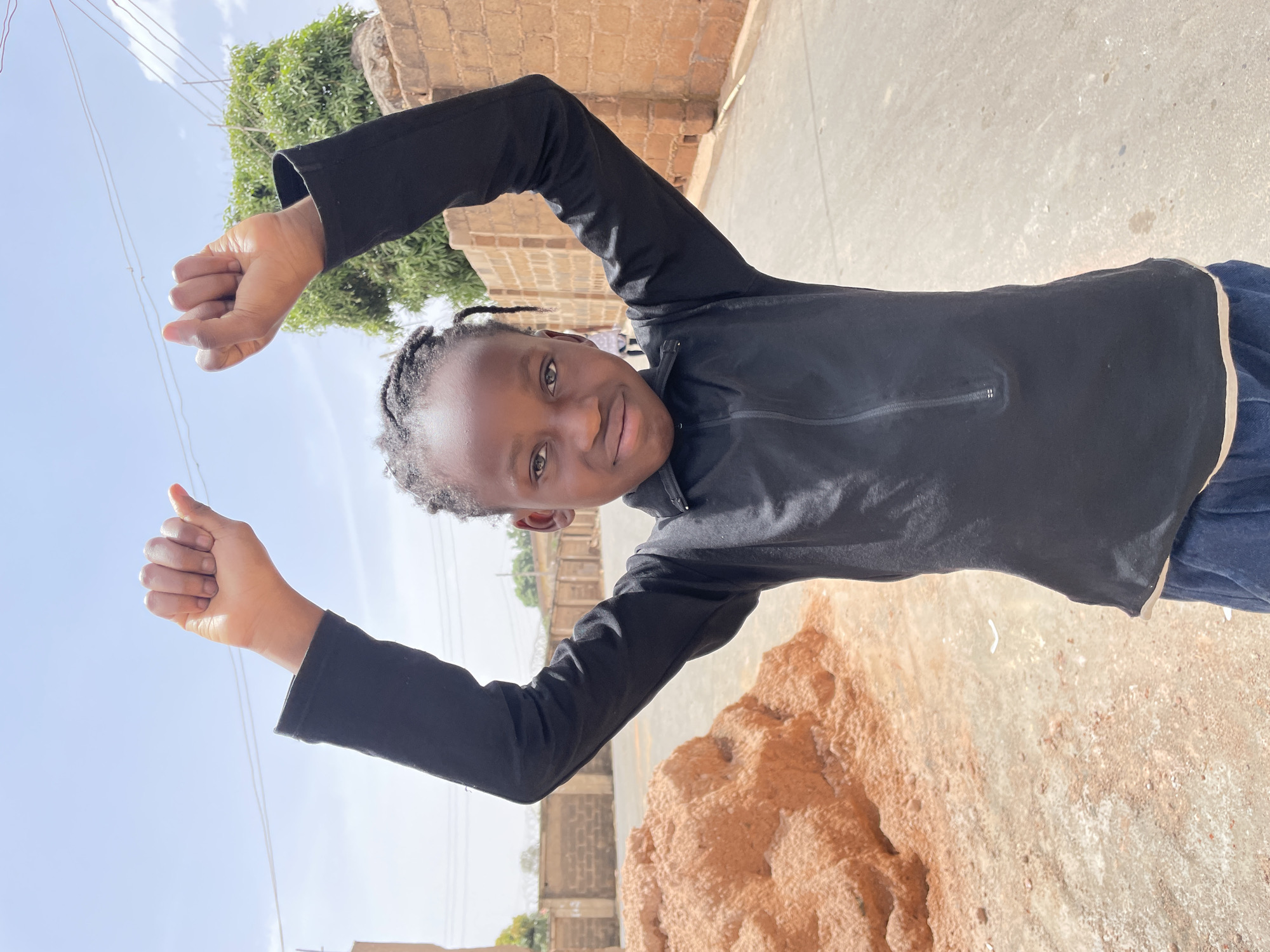 A young, dark-skinned child with braided hair stands facing forward on a mixed surface of light-colored pavement and reddish-brown earth, located in Jos, Nigeria. The child wears a black long-sleeved top with a quarter-zip neckline and dark blue pants. Both arms are raised above the head, with hands clenched into fists. The child exhibits a slight, closed-mouth smile while looking directly at the camera. To the left of the child, a substantial mound of reddish-brown soil is present. The background features a brick wall, partially topped with barbed wire, and green tree foliage on the right. Further left, a light-colored building structure and overhead utility wires are visible. The scene is illuminated by bright daylight under a pale blue sky with scattered wispy clouds.