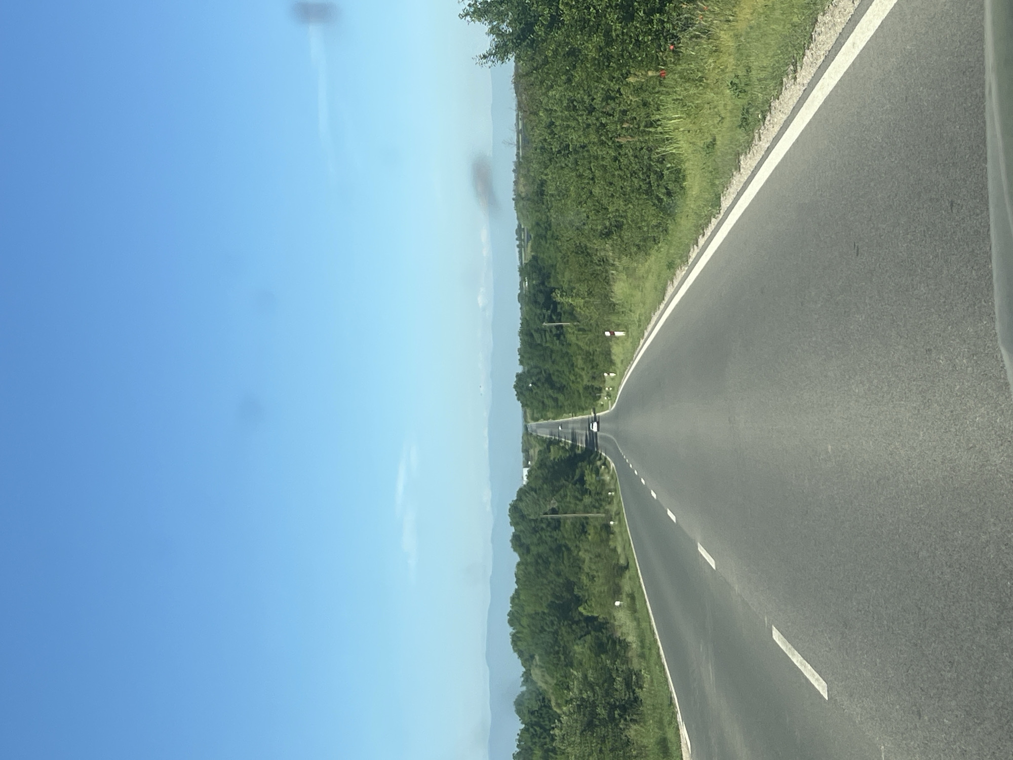 The image depicts a straight stretch of asphalt road in Brezovo, Bulgaria, flanked by green vegetation on both sides. A clear blue sky occupies the left portion of the frame. In the distance, the road narrows to a vanishing point, with other vehicles visible along the asphalt. Road markings, including dashed white lines, are visible. The vegetation alongside the road appears to be a mix of trees and grasses. Mountains are faintly visible on the horizon. The image appears to have been taken from within a vehicle.