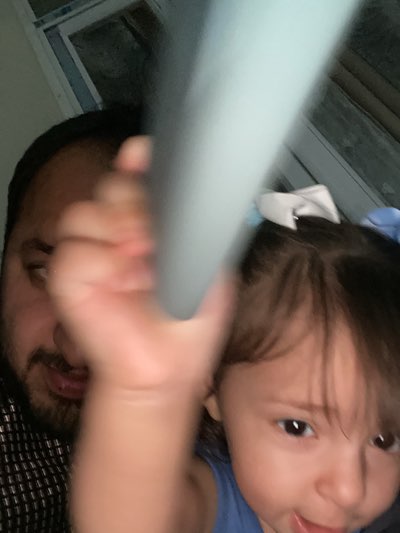 A man and a toddler are in the foreground, with the man's face partially visible on the left side of the image. He has dark hair and a beard. The toddler, wearing a blue shirt and hair bow, is on the right side, partially obscured by a blurry light blue object held by a hand. The background includes a window with a visible grid pattern. The location cannot be determined to be in Kandahar, Afghanistan, based solely on this image.