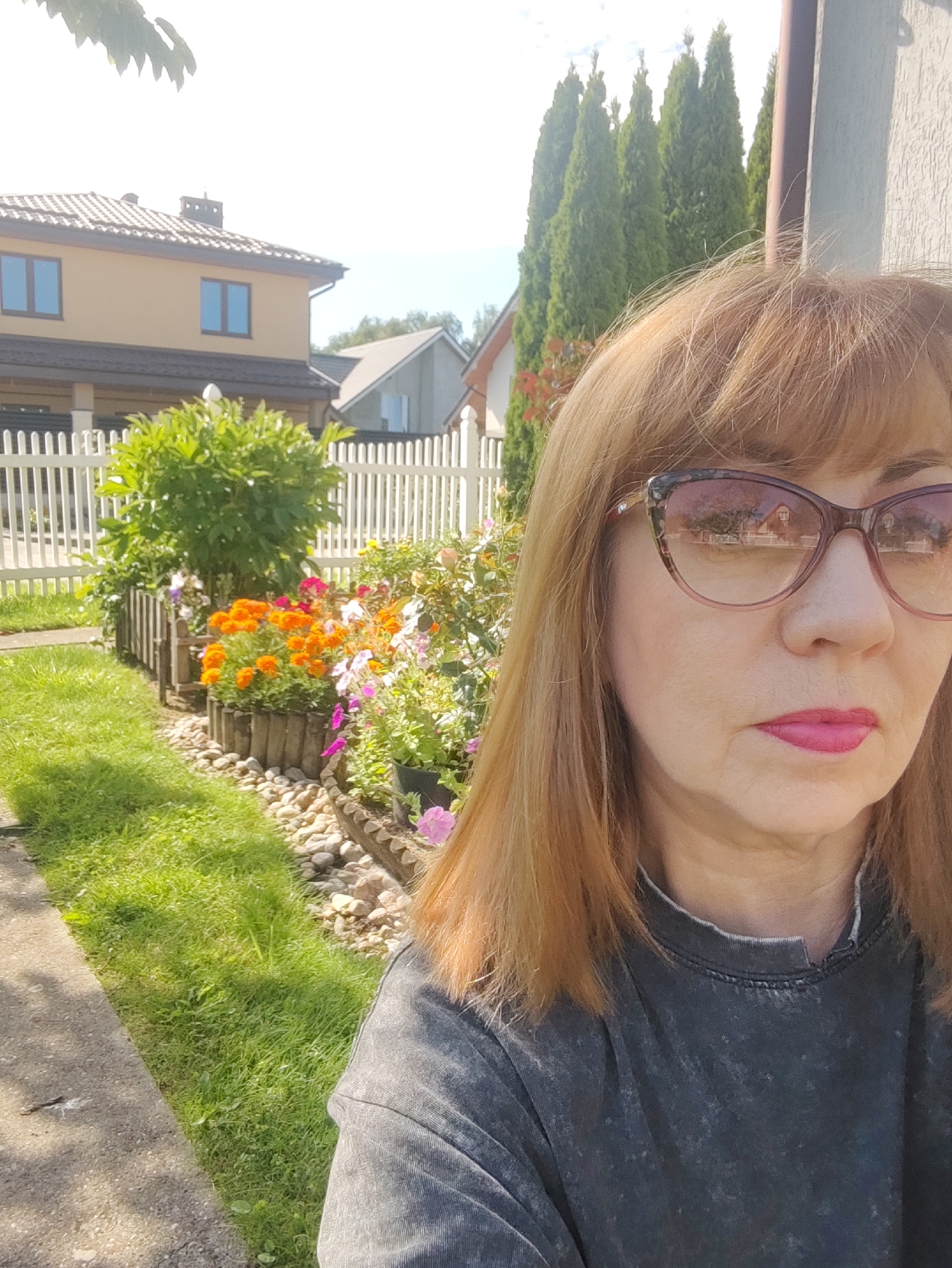 A woman with blonde hair and glasses takes a selfie in front of a garden with flowers and a white picket fence. Behind the garden are two houses and several tall green trees. The woman is wearing a dark gray t-shirt. The scene appears to be in a residential area on a sunny day. The image is not specific to Gvardeysk, Russia.