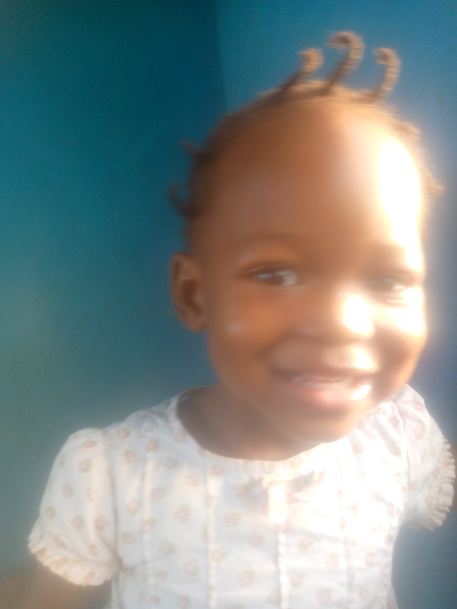 A young child is smiling at the camera. The child has dark skin and short, styled hair. The child is wearing a white shirt with a floral pattern. The background is a solid blue wall. The photograph is taken in a brightly lit environment, but the image appears to be slightly out of focus, obscuring finer details. The location in the city is Unknown, Unknown.