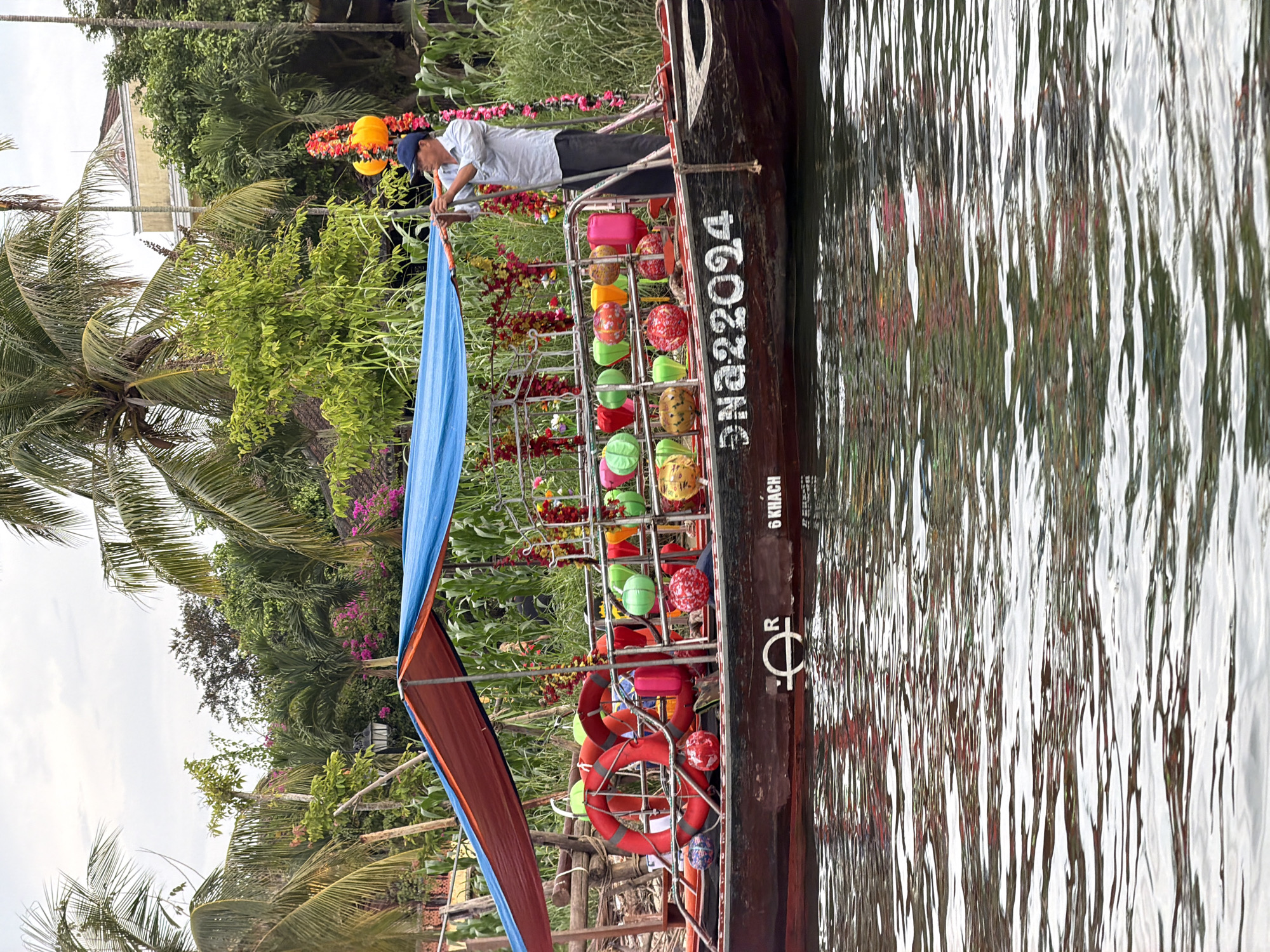 A traditional wooden boat is depicted on a calm body of water, likely a river or canal, amidst a lush tropical landscape. The scene is dominated by dense green foliage, including tall palm trees and various bushes, which line the banks.
On the boat, a person, appearing to be a man, is visible standing near the front. He wears a light-colored collared shirt and a blue cap, and is engaged in what looks like adjusting a part of the boat's structure or canopy.
The boat itself is dark-stained wood, featuring a canopy at the bow with blue and reddish-orange fabric panels. Along its side, a metal rack holds numerous colorful spherical decorations, resembling lanterns or patterned globes, in hues of red, yellow, orange, green, and blue. Several red life rings are also hanging on the boat's side. The water reflects the surrounding greenery and sky, showing subtle ripples.
In the background, partially obscured by the foliage, a light-colored building with architectural details indicative of local design can be seen. The sky is overcast, suggesting a daytime scene under cloudy conditions.
Visible text on the side of the boat includes "DN.A 22024," which appears to be a registration number. Below this, the words "6 KHACH" are clearly legible, translating from Vietnamese to "6 guests" or "6 passengers," indicating the boat's capacity. A distinctive symbol resembling a stylized 'R' or a currency mark is also present on the boat's hull.
The image captures a peaceful moment on a sightseeing or transport boat, characteristic of the waterways in places like Hội An, Vietnam.