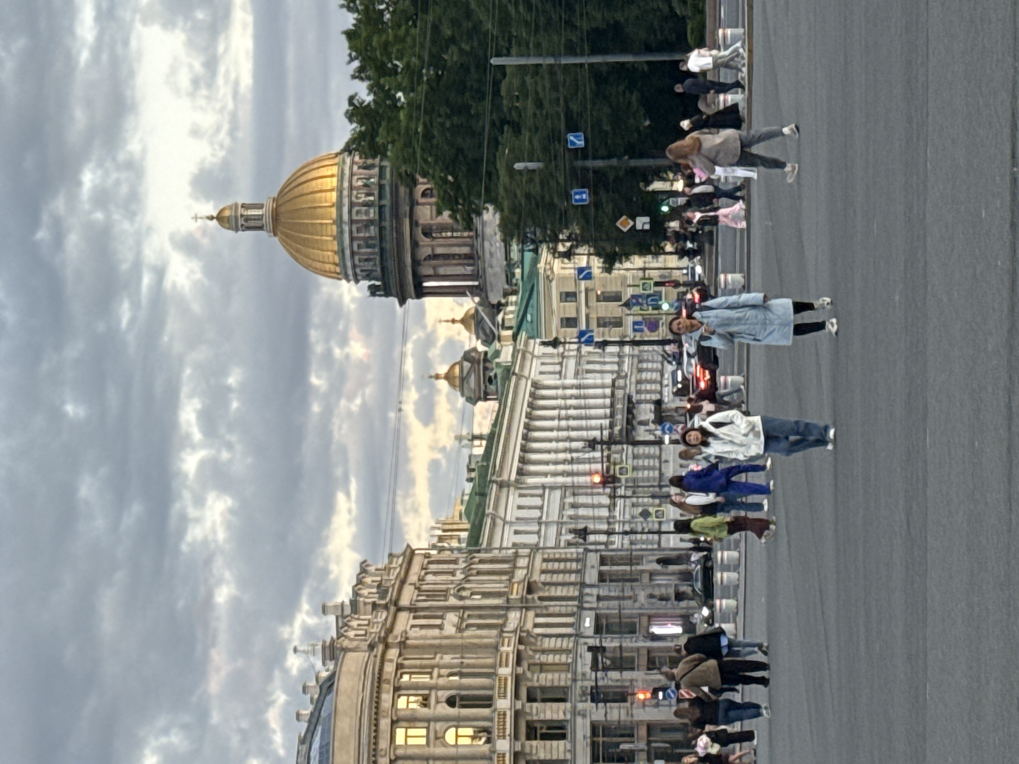 This is a photograph taken at Palace Square in Saint Petersburg, Russia, with Saint Isaac's Cathedral prominently visible in the background. The sky is overcast with diffused light, suggesting either early morning or late evening.
In the foreground, a pedestrian crossing occupies the lower portion of the frame. Several individuals are walking across the square. Closest to the viewer, a woman wearing a light blue coat and dark pants walks away from the camera. To her right, slightly further back, another woman in a white jacket and blue jeans is walking towards the camera. A diverse group of people, some walking in pairs or small groups, are distributed across the square, engaged in transit.
The middle ground features a collection of grand, classical buildings that line Palace Square. These structures exhibit intricate architectural details with multiple stories, columns, and ornate facades. Traffic lights and street signs are visible, indicating a functional urban intersection. Several cars are present, suggesting ongoing city traffic.
In the background, the golden dome of Saint Isaac's Cathedral dominates the skyline, its grandeur amplified by the overcast sky. Smaller domes of other buildings can be seen to the left and right of the main cathedral. Trees are present, forming a green buffer between the buildings and the sky. The overall scene depicts a bustling urban environment with a focus on historical architecture and everyday pedestrian activity.