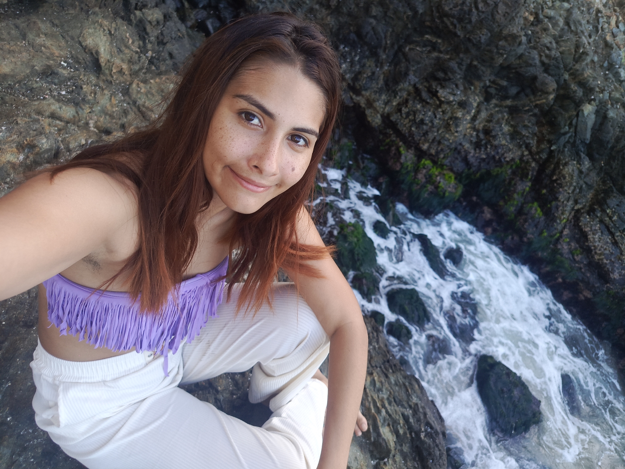 In the city of Pedro González, Venezuela, a young woman with reddish-brown hair and freckles is captured in a selfie. She is seated on rocky terrain next to the ocean. She is wearing a light purple bikini top with fringe detailing and loose-fitting white pants. The background consists of dark, wet rocks and the turbulent ocean water.
