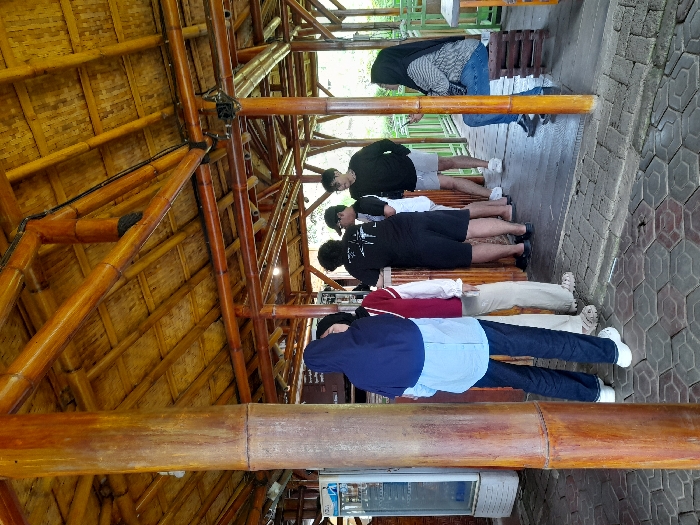 The image depicts a scene inside a bamboo-constructed structure, likely a restaurant or cafe. The roof is made of woven bamboo panels, while the supporting beams are made of larger bamboo sections. Five people are present, including four adults and one child. Three adults stand in a line, facing a counter, presumably placing orders. A young woman wearing a blue shirt, jeans, and a black headscarf stands at the front of the line, while a woman in a red shirt and beige pants stands behind her, and a man wearing a black t-shirt and shorts stands behind her. A young man wearing a white shirt, dark pants, and a blue baseball cap appears to be speaking with the man in black. To the right, a woman wearing a black headscarf and a blue denim jacket sits on a wooden bench, holding a phone in her hand. The floor is covered in grey interlocking pavers. The location is a cafe in Kecamatan Pujon, Indonesia, at the coordinates -7.854 latitude and 112.453 longitude.
