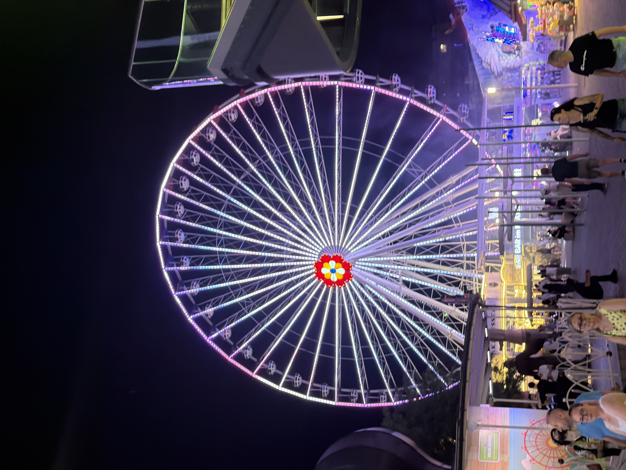 The image depicts a night scene at the Prater amusement park in Vienna, Austria. The main subject is the Wiener Riesenrad, a large Ferris wheel illuminated with colorful LED lights, predominantly white, pink, and blue. The Ferris wheel has a distinctive red, yellow, and blue flower-like decoration at its center. In the foreground and midground, various people are walking and standing in a paved area. Behind the Ferris wheel, there are amusement park structures with bright lights, including what appears to be a building with signage that reads "BAR im." The overall atmosphere is festive and characteristic of an amusement park at night.