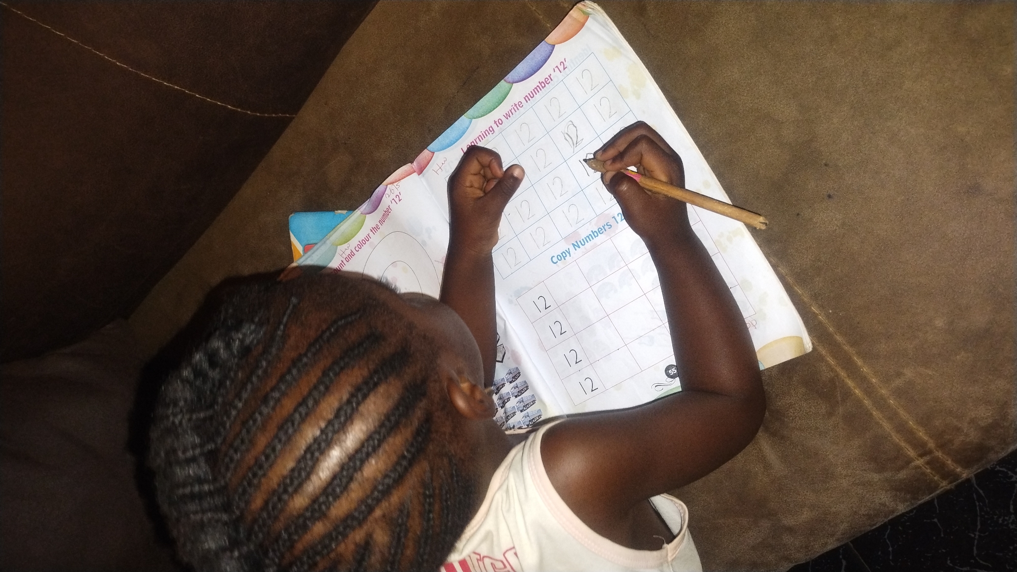 A young child, likely a girl due to her braided hairstyle, is sitting on a brown couch, writing the number "12" in a workbook. The workbook page is titled "Learning to write number '12'" and has pre-written examples and space to copy. The child is using a wooden pencil. The photo was taken in Jos, Nigeria.