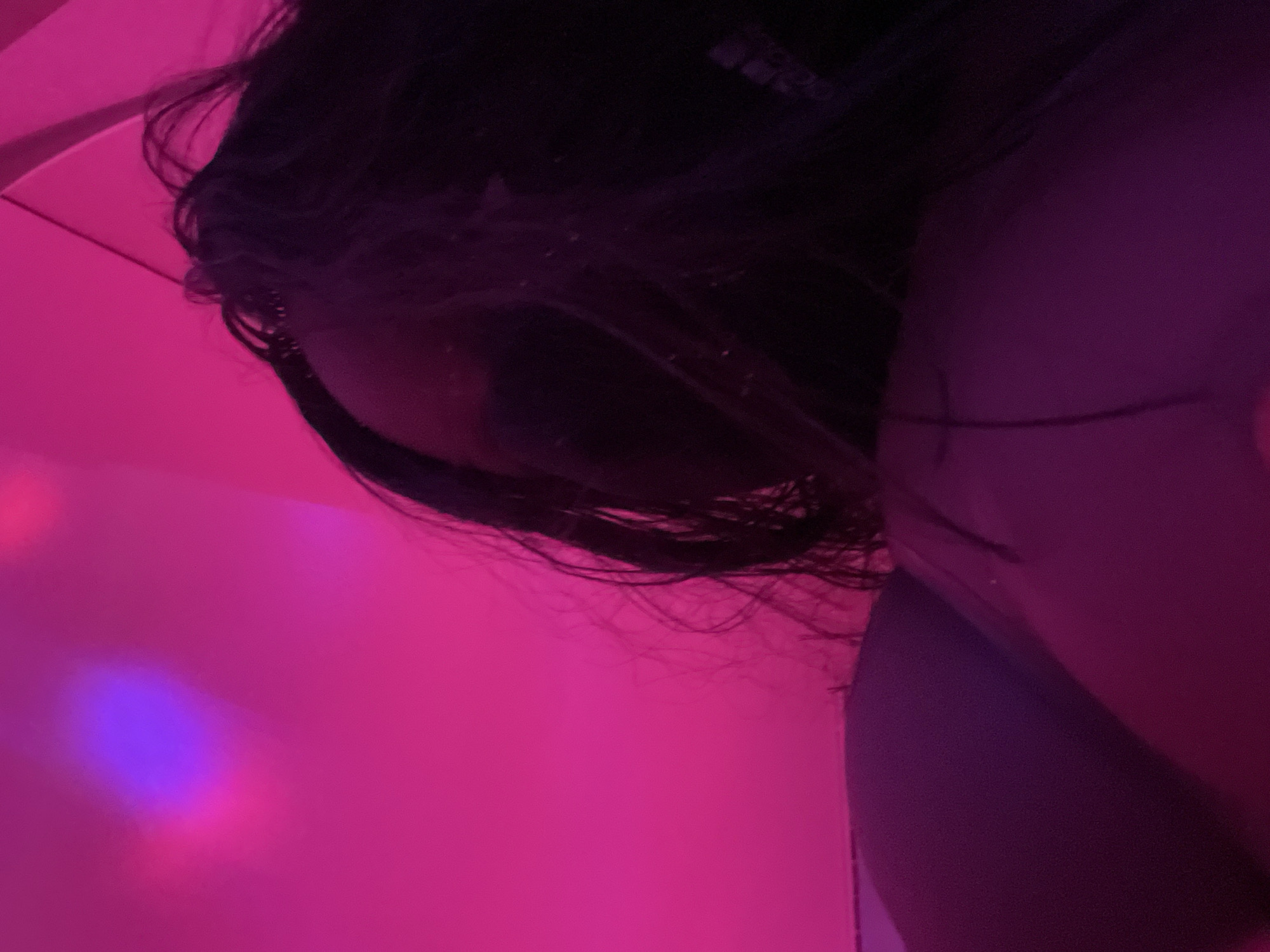 In a dimly lit room with a vibrant pink hue, a person's dark hair is partially visible, cascading down and obscuring their face. The lighting casts an intense, saturated glow on the surroundings, creating a hazy and dreamlike atmosphere. The image is a close-up, focusing on the hair and a portion of the person's body, suggesting an intimate or personal moment captured in time. The background features a gradient of pink and purple, with subtle hints of blue light.