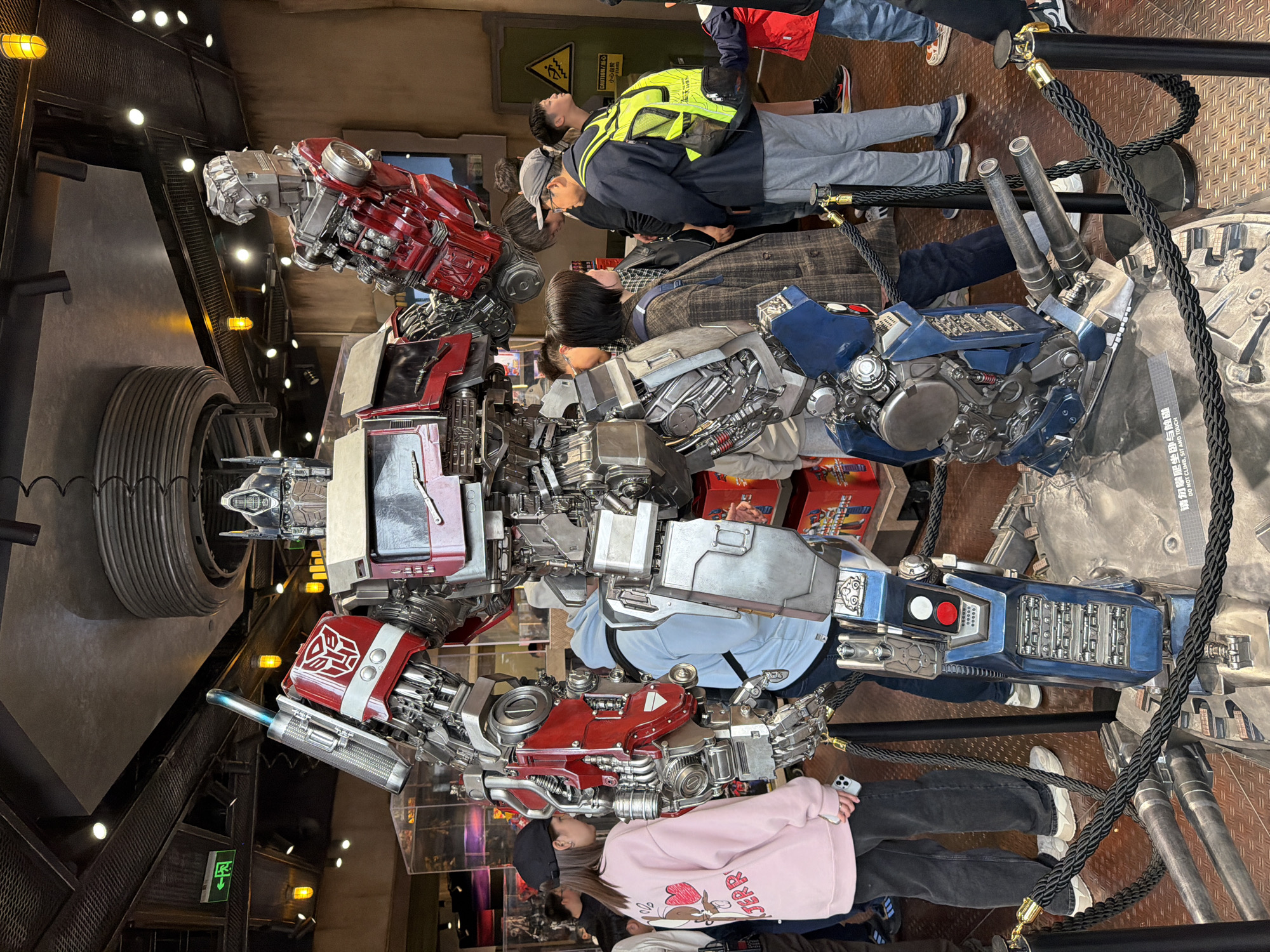 The image shows a full-body statue of a Transformer robot in a public setting. People are standing around and near the statue, some taking pictures. The statue is predominantly red, silver, and blue, with intricate mechanical detailing. It is cordoned off by black ropes supported by metal poles, likely to keep the public at a safe distance. Signs are visible in the background that include Chinese characters and English phrases like "DO NOT CLIMB, SIT AND TOUCH" and "CAUTION/小心台阶", suggesting that the scene is in Tongzhou District, China.