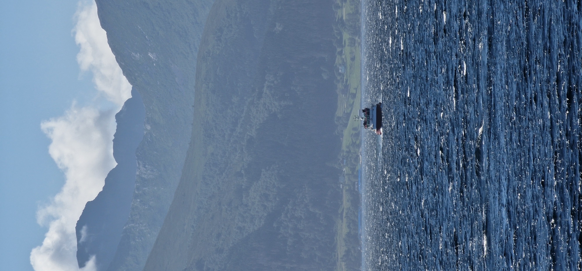 A medium-sized, white and red boat with several people visible on its deck, travels across a choppy, dark blue body of water. In the distance, a densely forested mountainside slopes down to a line of buildings and green fields. The background is dominated by hazy, layered mountain ranges under a pale blue sky with scattered white clouds. This scene is likely located in the vicinity of Volda, Norway, a town known for its dramatic fjords and mountainous terrain.