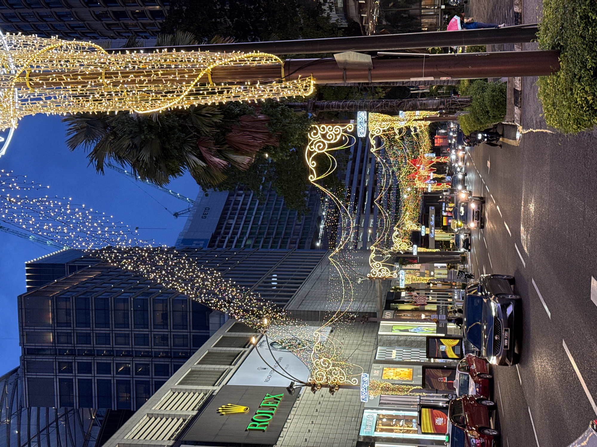 The image depicts a street in Kuala Lumpur, Malaysia at night, adorned with festive lights. A street with traffic is surrounded by buildings. Along the street are decorations of illuminated lights with swirly, decorative features. Storefronts, including one with a prominent "ROLEX" sign, line the street. Vehicles, including cars and motorcycles, are seen driving along the wet road. A crane is visible in the background, indicating construction activity. On the sidewalk, a person is visible, lying on their back.