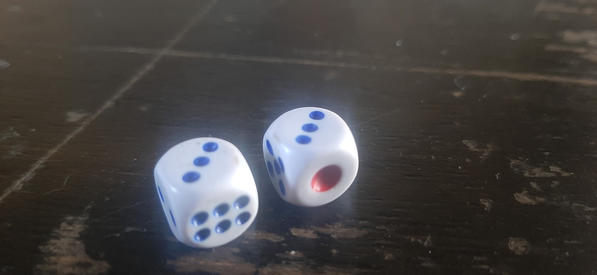 Two white dice with blue pips rest on a dark, textured surface that appears to be a wooden table with visible grain and faint scratches. The die on the left shows faces with three pips and six pips. The die on the right displays faces with three pips and five pips, and a red-colored pip on one of its visible sides. The lighting suggests an indoor setting with a slight shadow cast by the dice. The background is dark and out of focus, with faint diagonal lines that may indicate the surface is a tiled or paneled area. The scene is devoid of any human presence or activity. No information about the location in Yessentukskaya, Russia can be derived from the visual elements of the image.